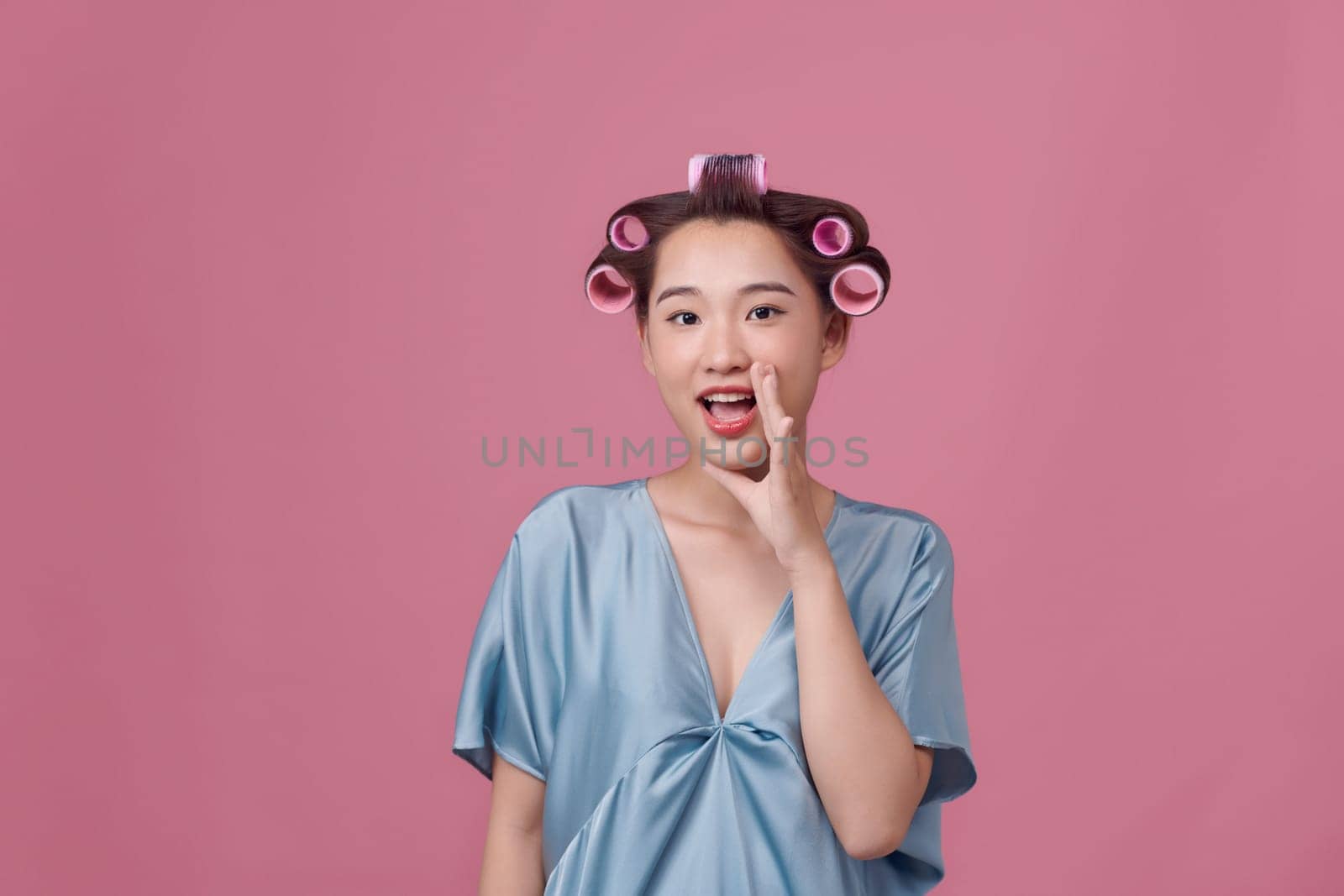 Happy young woman in blue dress and hair curlers on pink background by makidotvn