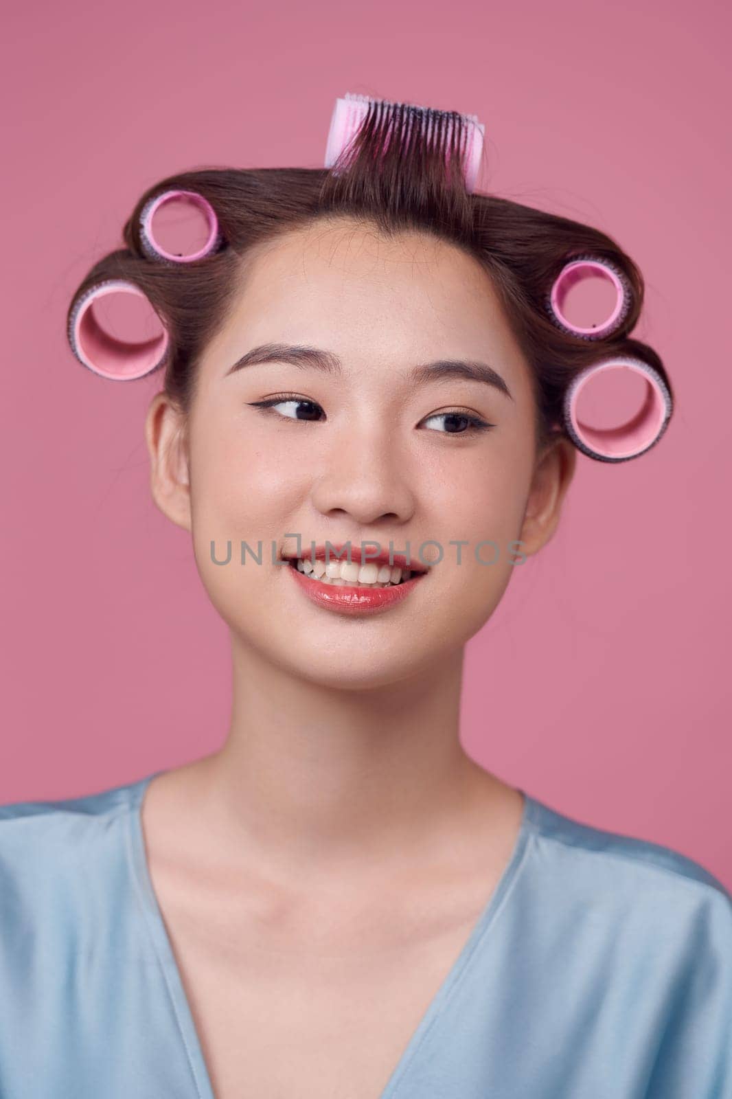 Happy young woman in blue dress and hair curlers on pink background