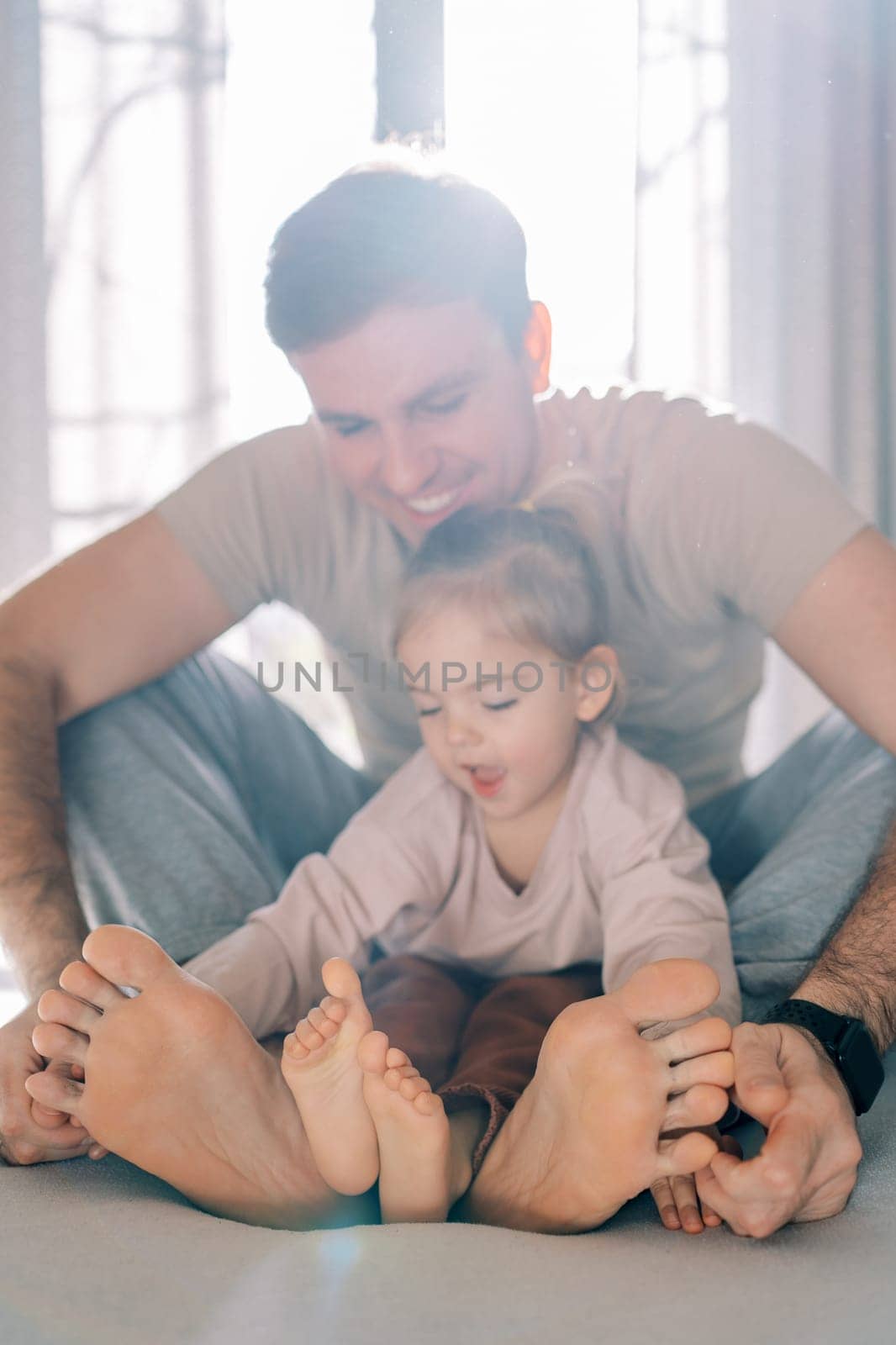 Little girl sits with her dad on the bed and examines their bare feet by Nadtochiy