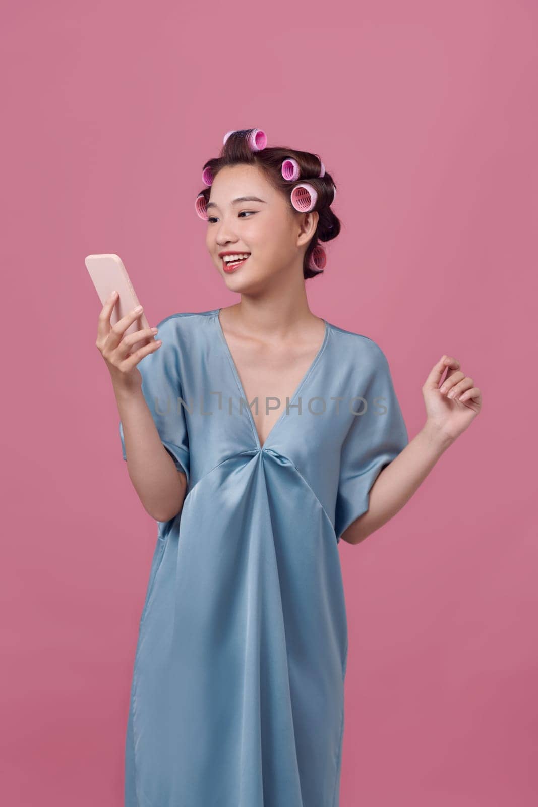 Pretty asian woman in hair rollers using smartphone on pink background by makidotvn