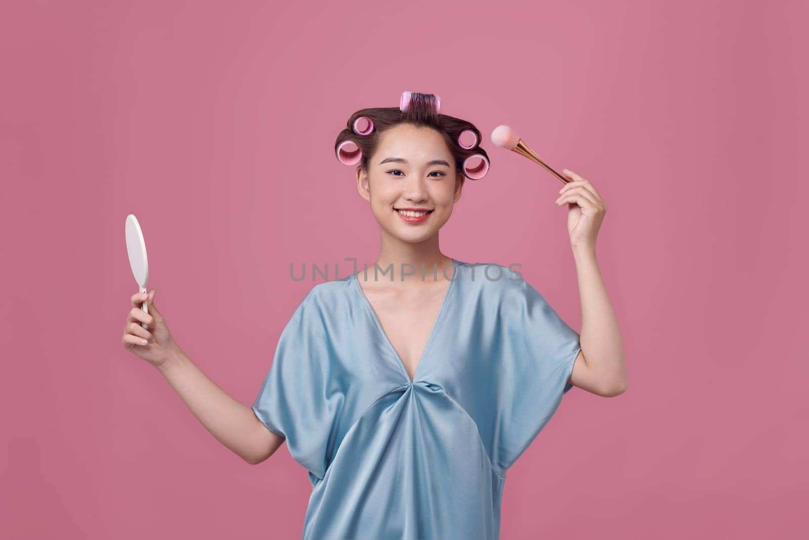 Pretty woman with curlers on hair, holding makeup brushes and mirror in hands 