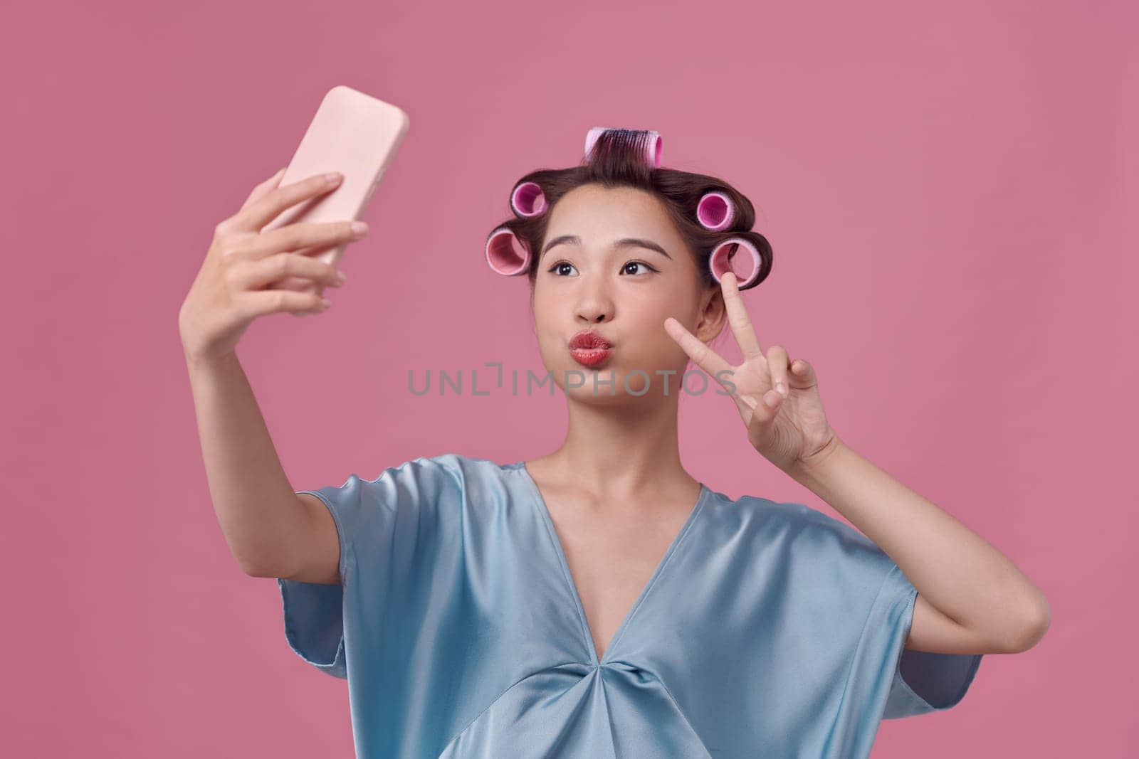 cheerful woman applies hair curlers posing in smartphone camera dressed over pink background. by makidotvn
