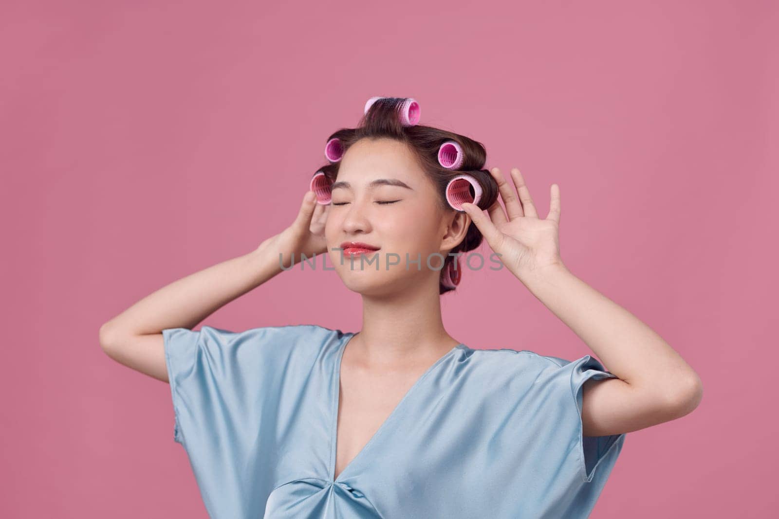 young beautiful girl having hair curlers on her head isolated on pink background