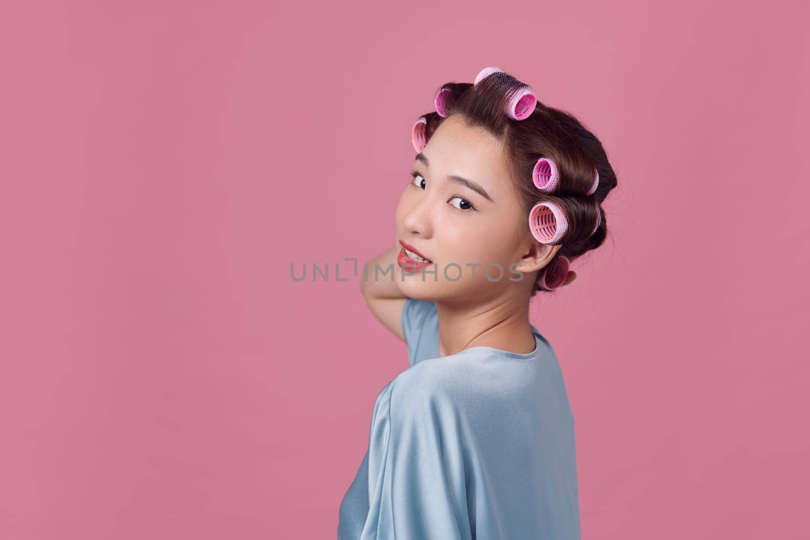 unsure and smiling young woman without makeup and with hair curlers on pink