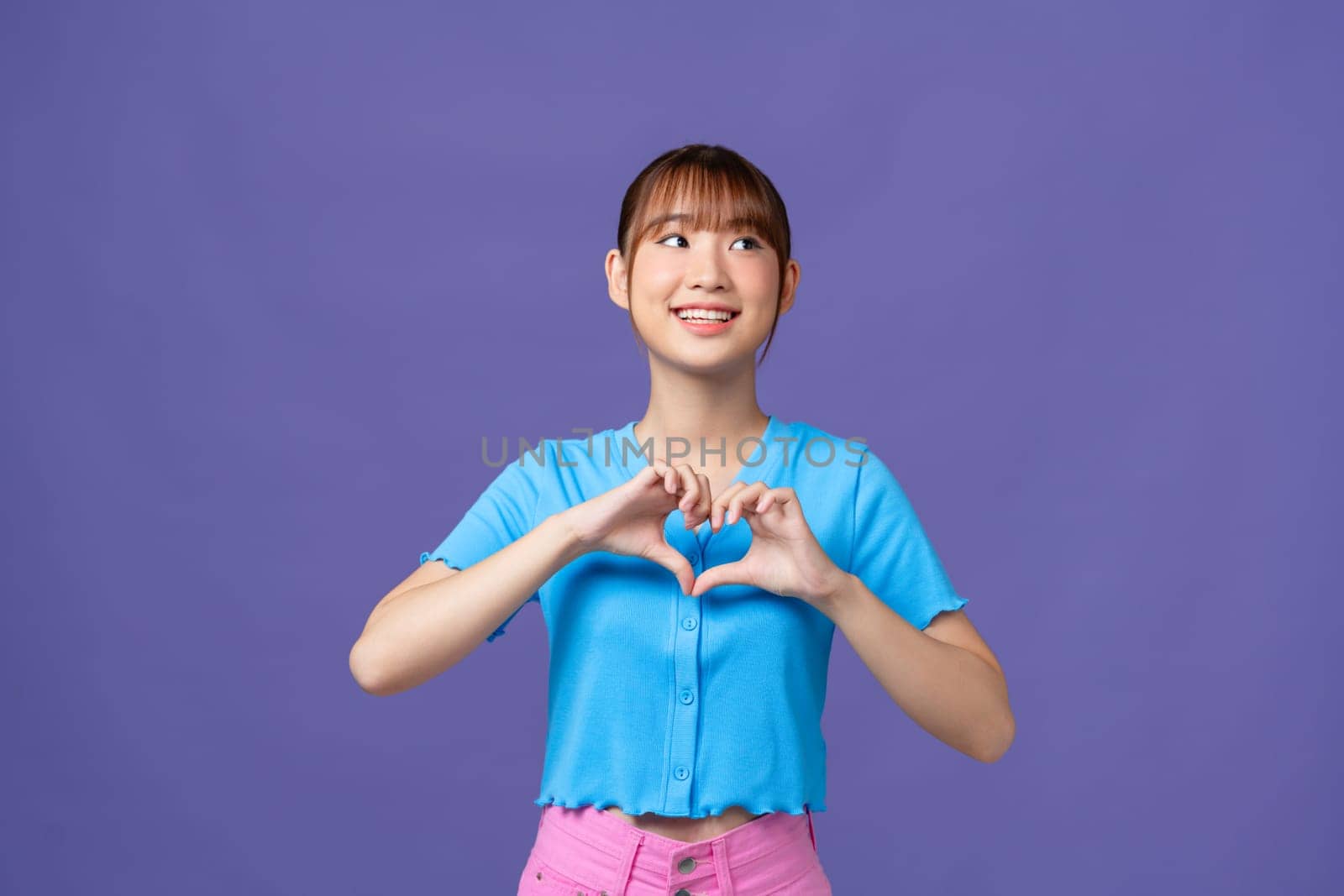 Young asian woman shows her fingers heart-shaped sign and presses to chest on purple background. by makidotvn