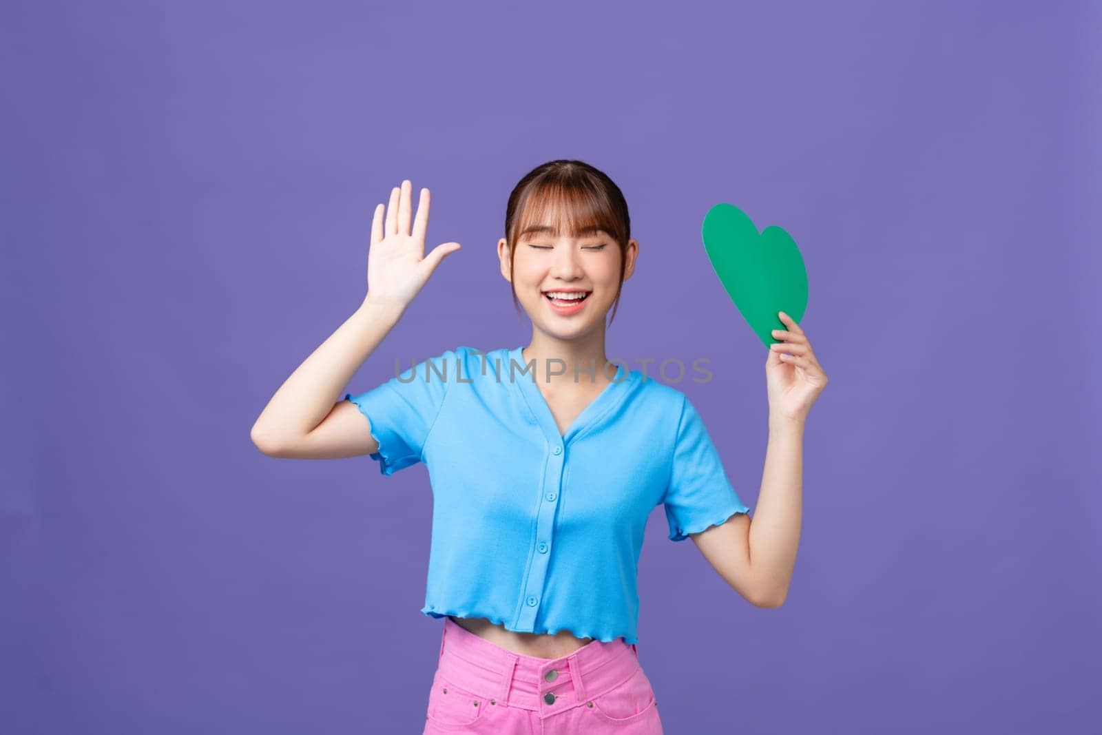 Sweet adorable lady wear blue crop top holding green heart isolated purple background by makidotvn