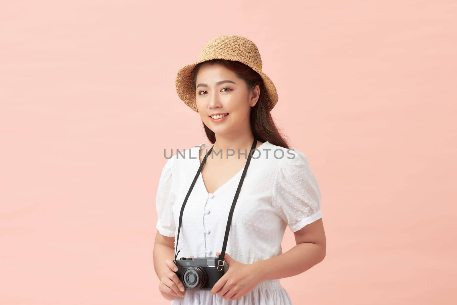 A smiling young woman in summer hat standing with photo camera isolated over pink background