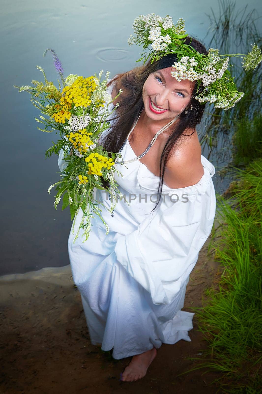 Adult mature brunette woman in a white dress, sundress and a wreath of flowers in summer by the water of river or lake in the evening at sunset. Celebration of the Slavic pagan holiday of Ivan Kupala by keleny