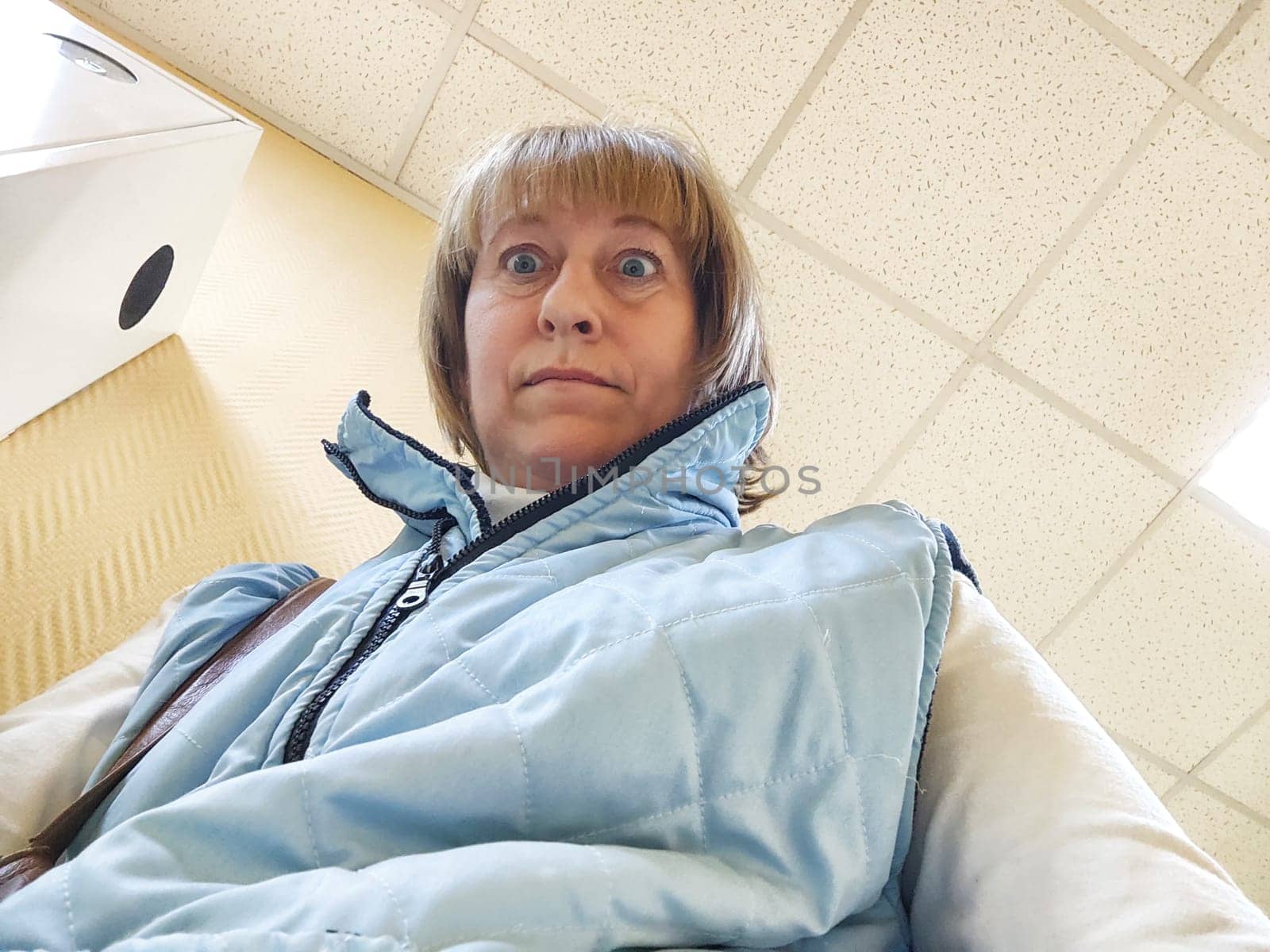 A strange girl poses against the background of a white ceiling with tiles. Funny middle-aged woman takes selfie with an unusual facial expression