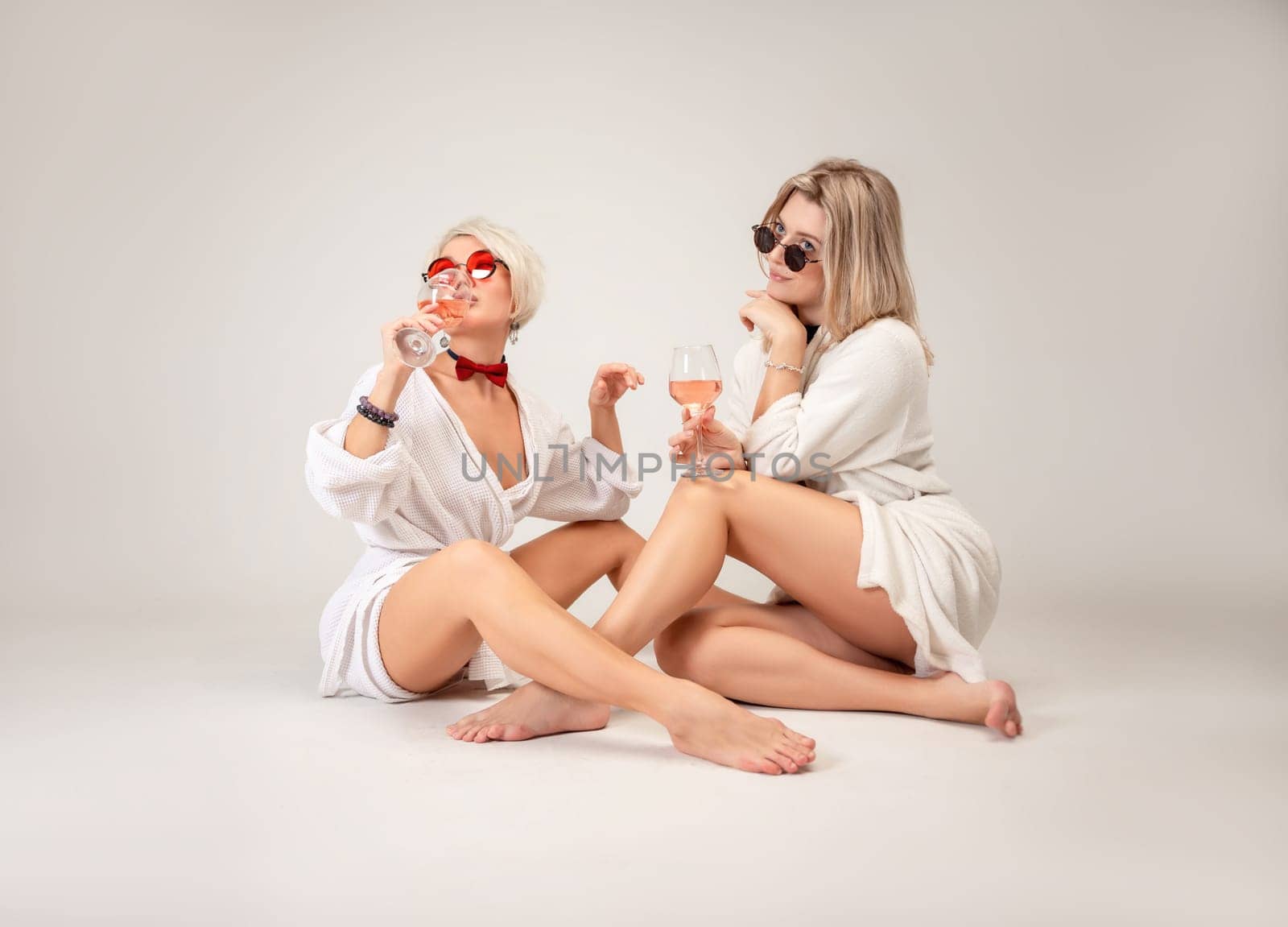 two girls in white coats and glasses with glasses of alcohol are sitting in the studio on a white background of a copy paste by Rotozey