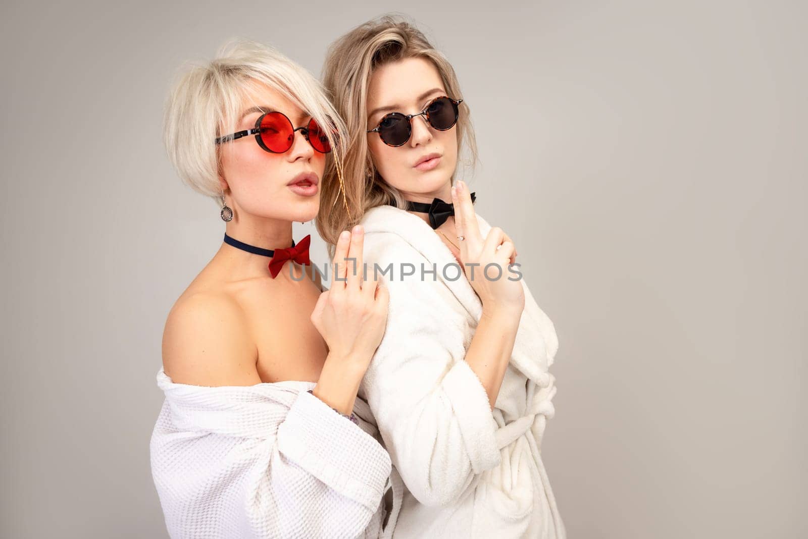 two close friends in white bathrobes and sunglasses stand close to each other and hold their fingers in the shape of a pistol