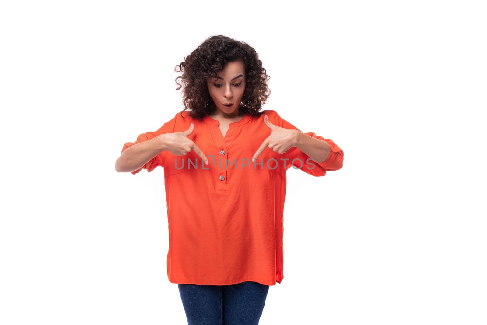 portrait of a bright young caucasian brunette woman with curly hair dressed in an orange shirt pointing her hand in surprise.