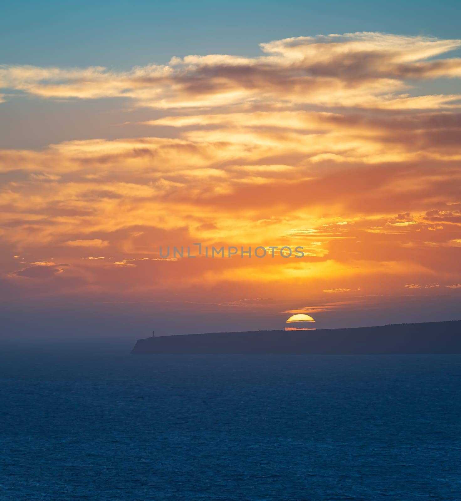 Spectacular sunset with clouds over cliffs and ocean, capped by a sentinel lighthouse.