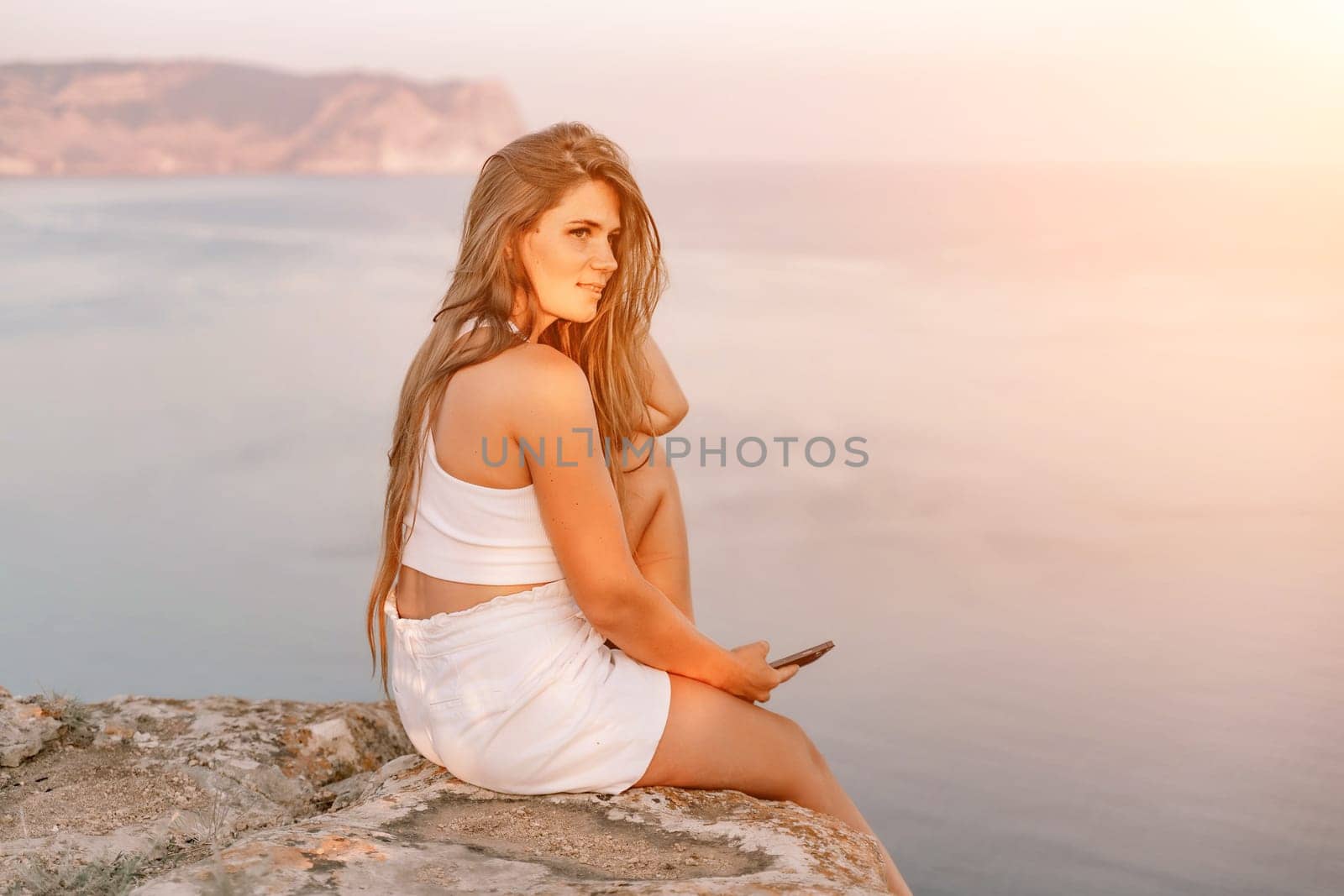Happy woman in white shorts and T-shirt, with long hair, talking on the phone while enjoying the scenic view of the sea in the background