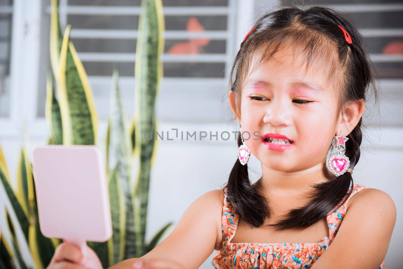 Asian adorable funny little girl making makeup her face and looking face herself in the mirror, Learning activity to be woman, happy kid is beautiful make up with cosmetics toy