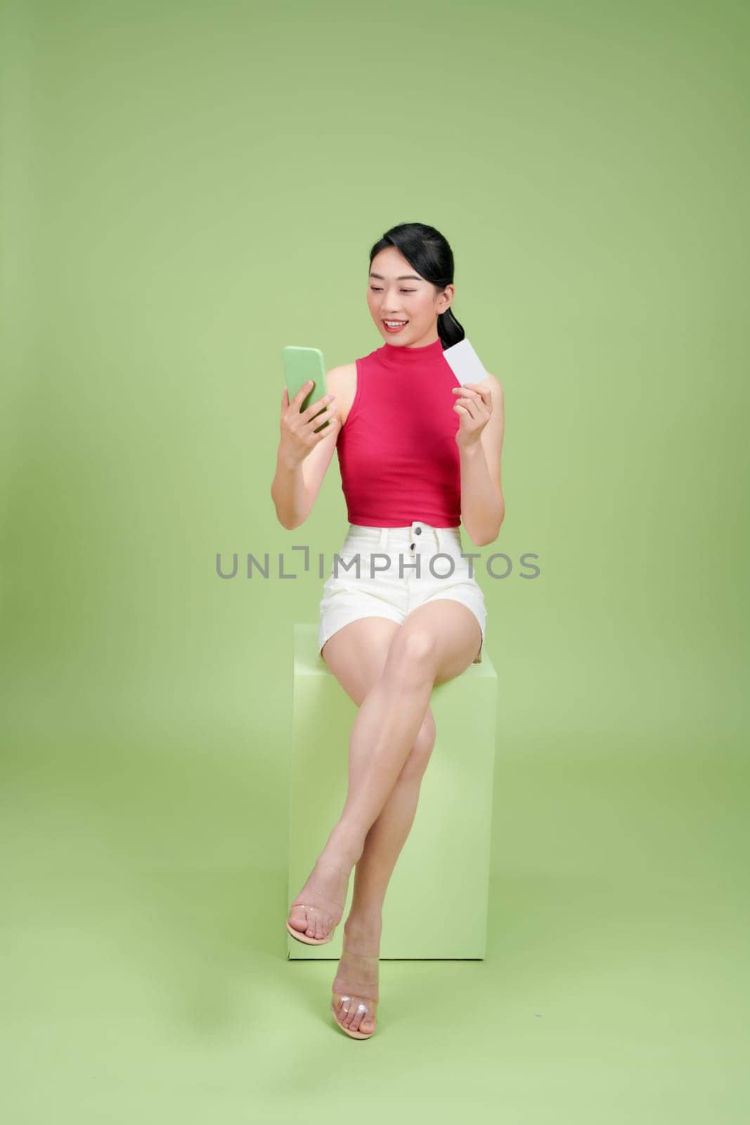 Young Asian businesswoman sitting on box and showing credit card and mobile phone isolated on green by makidotvn