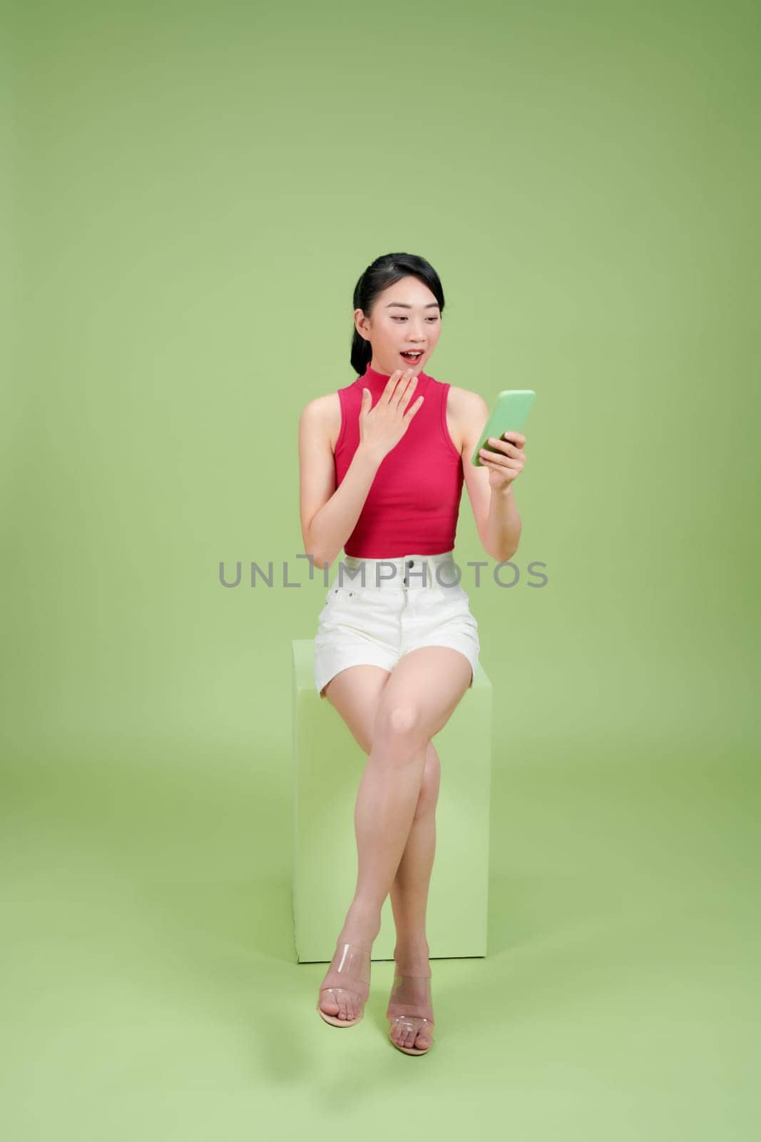 happy surprised young woman sitting using her mobile isolated on green background