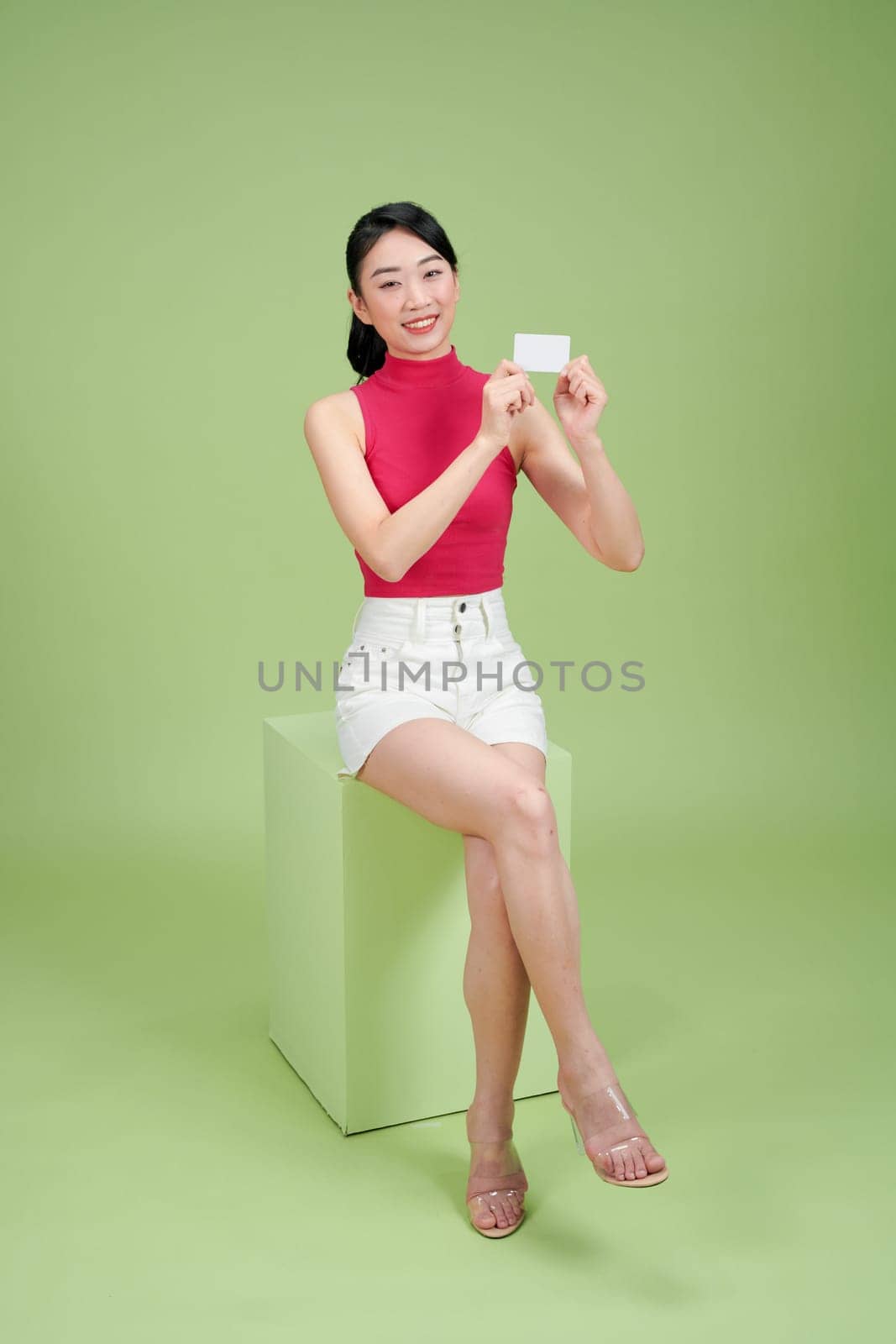 Attractive Asian girl laughing and holding credit card while sitting on podium over green background