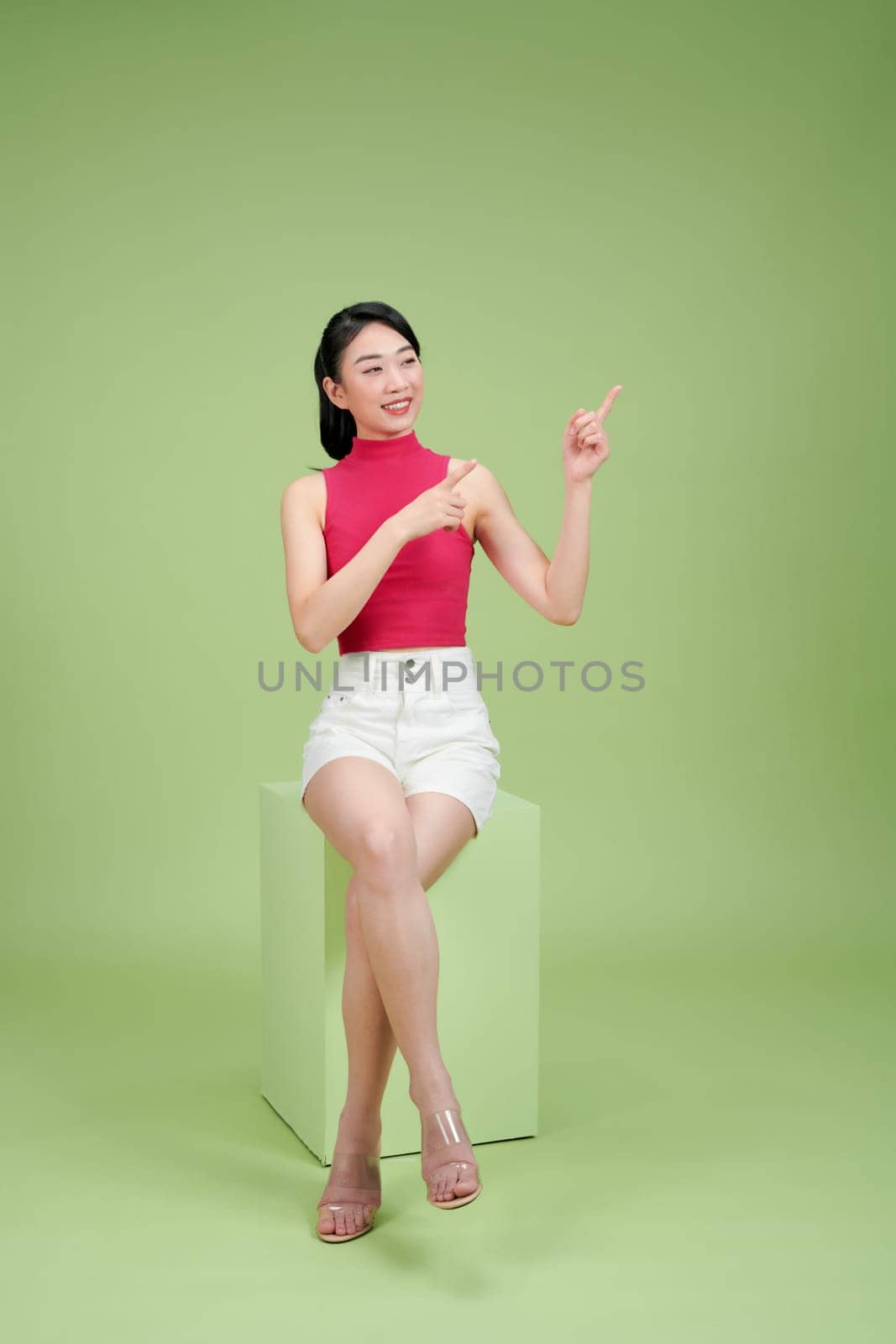 Happy young Asian woman sitting on podium and pointing finger up isolated on green background