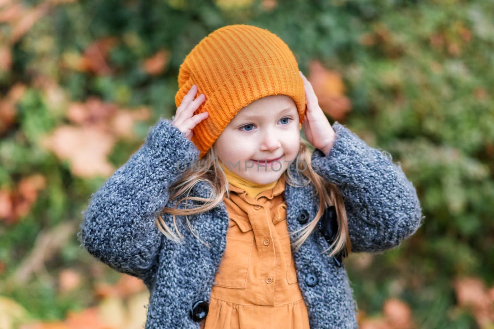 A little girl in an orange hat and a gray coat holds in a park in the autumn. by Godi