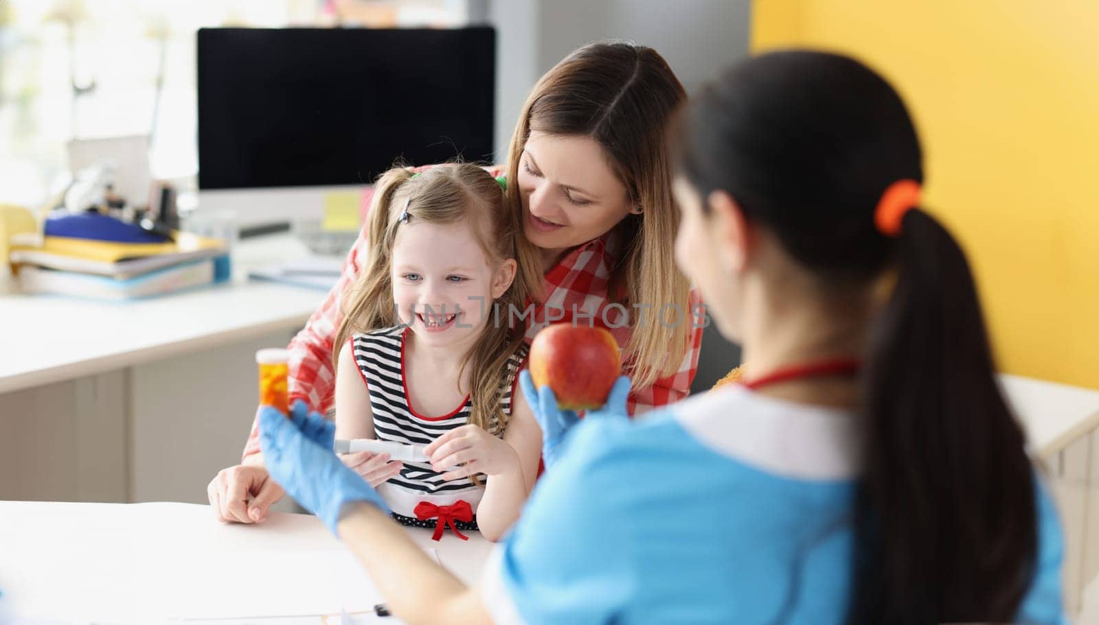 Doctor pediatrician holding bottle with medical capsules and red apple in front of little girl and mom in clinic. Vitamin complex for children concept