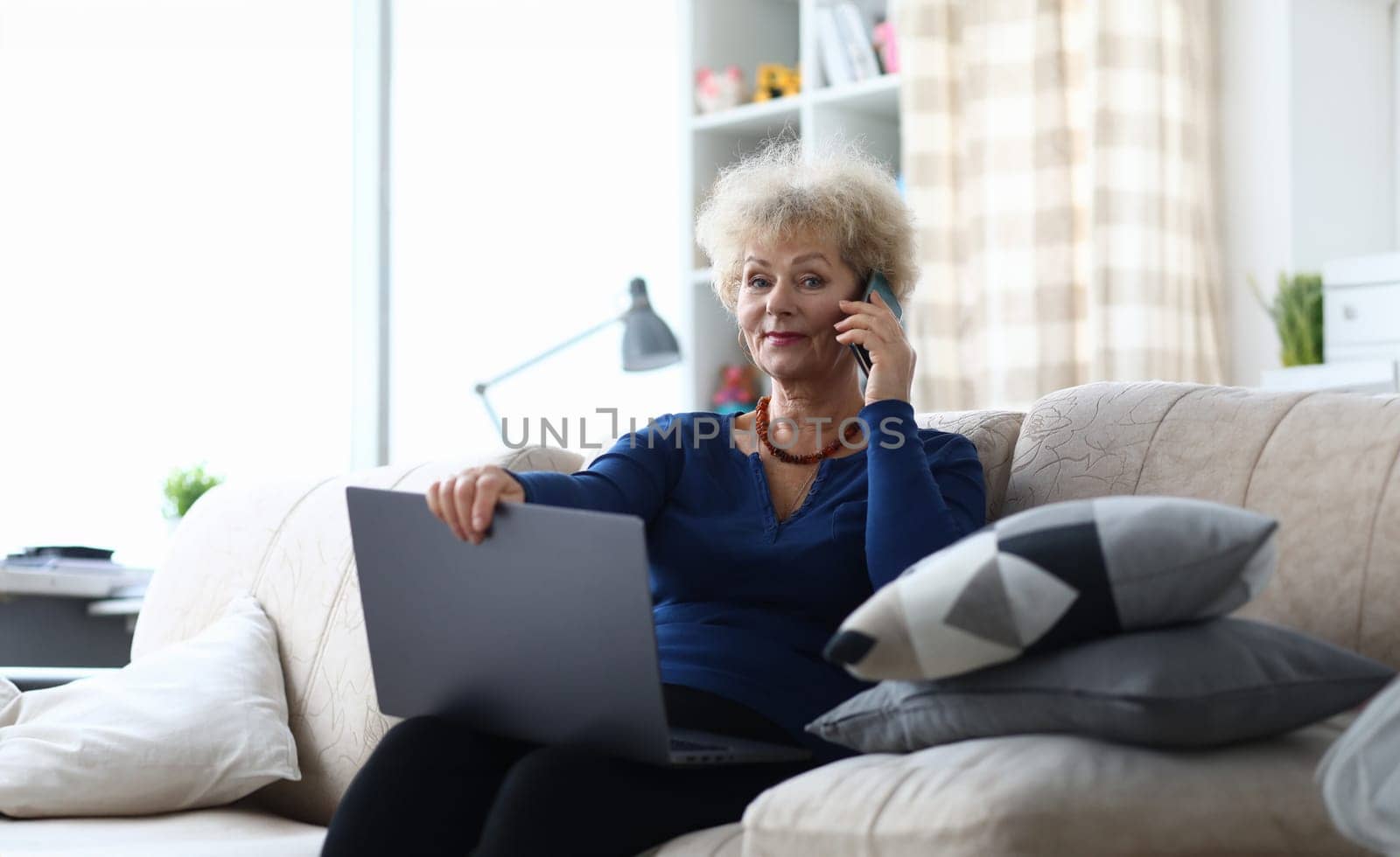 Portrait of beautiful elderly woman talking on mobile phone indoors. Businesswoman holding modern laptop and sitting on cozy couch in living room. Interesting communication. Technology concept