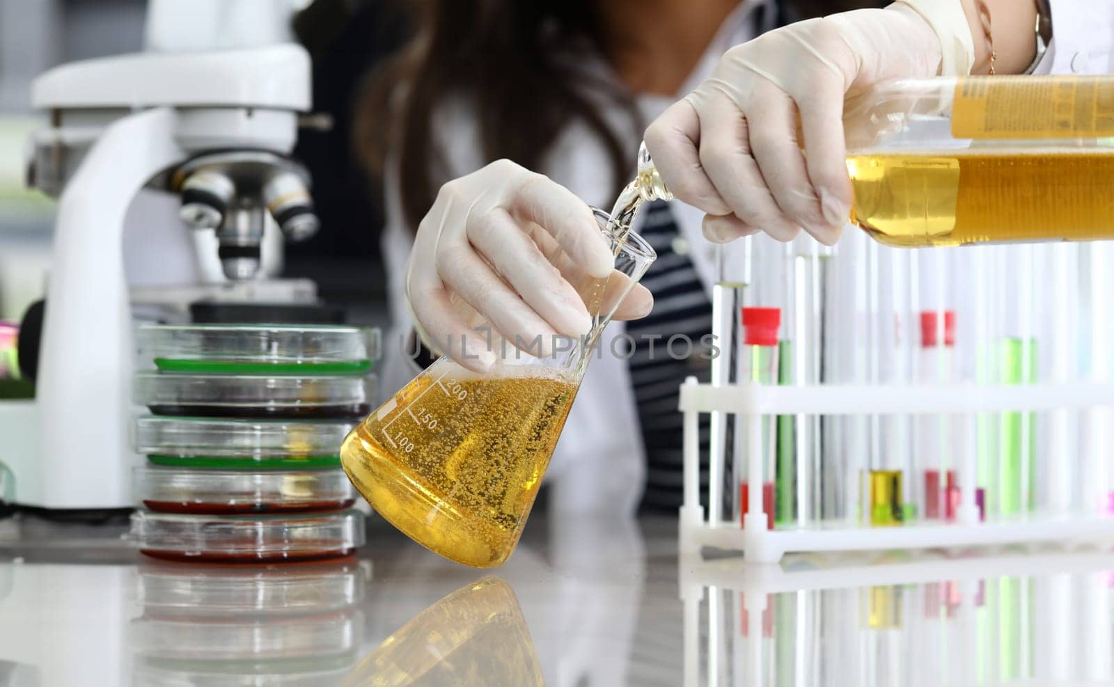 Close-up of laboratory female worker pouring alcohol liquid in glass flask. Woman working in white protective gloves and uniform. Chemistry and research. Modern investigation and science concept