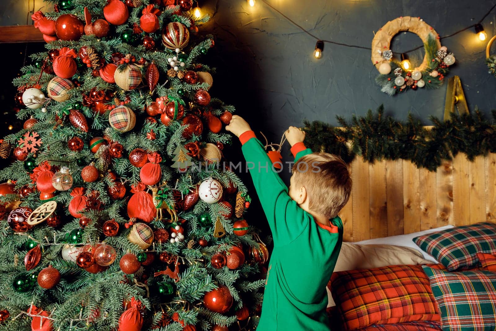 unrecognizable Child hanging a ball on illuminated Christmas tree.glowing lights. Boy in green pajamas decorating Christmas tree.Christmas good mood. Lifestyle, family and holiday. by YuliaYaspe1979