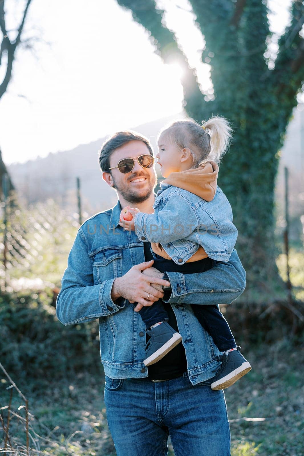 Smiling dad holding little girl with apple in his arms by Nadtochiy