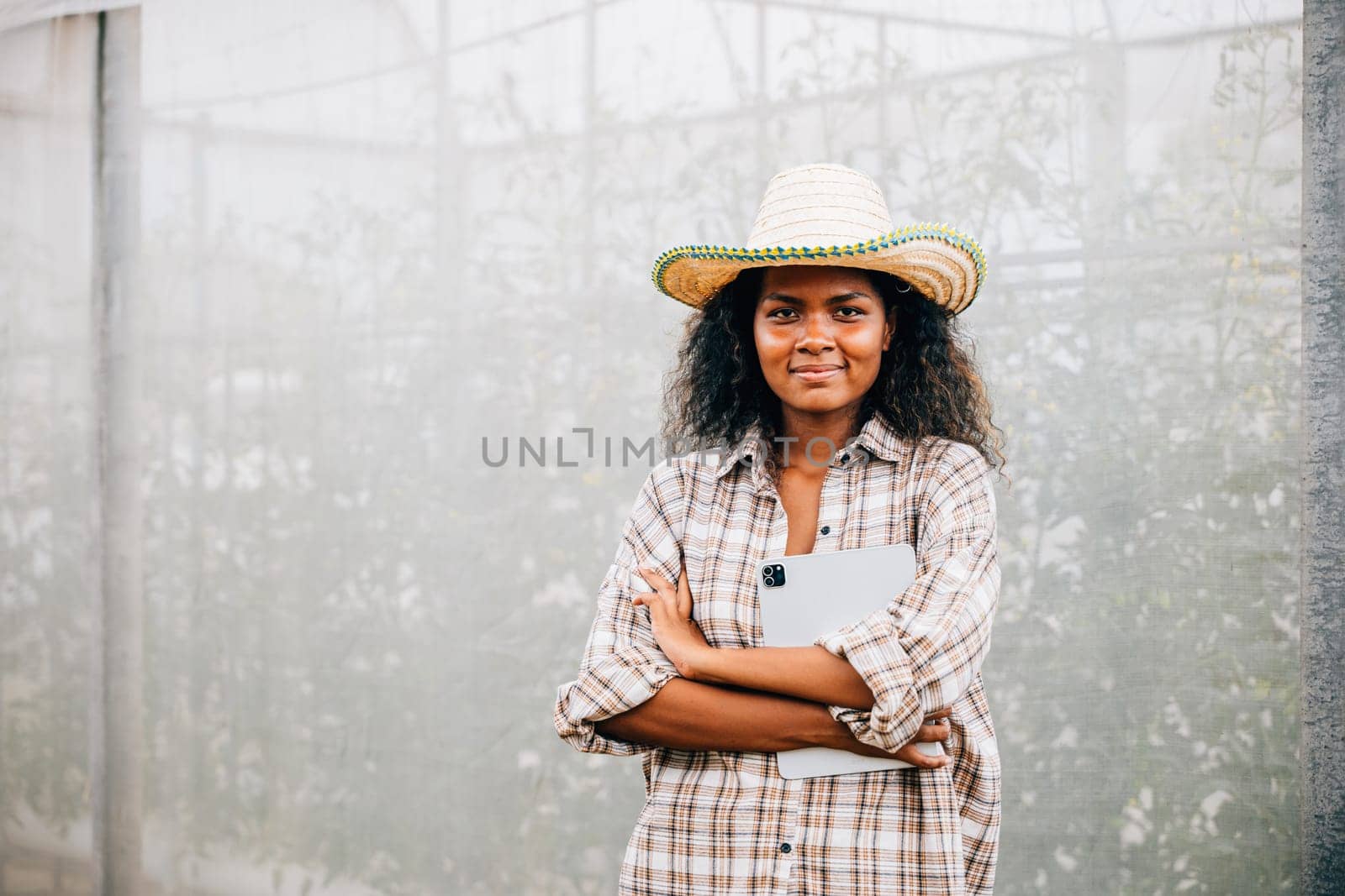 PhotoSuccessful greenhouse owner a young woman in a checked shirt and apron stands with arms crossed smiling of a woman posing confidently with a straw hat and crossed arms by Sorapop