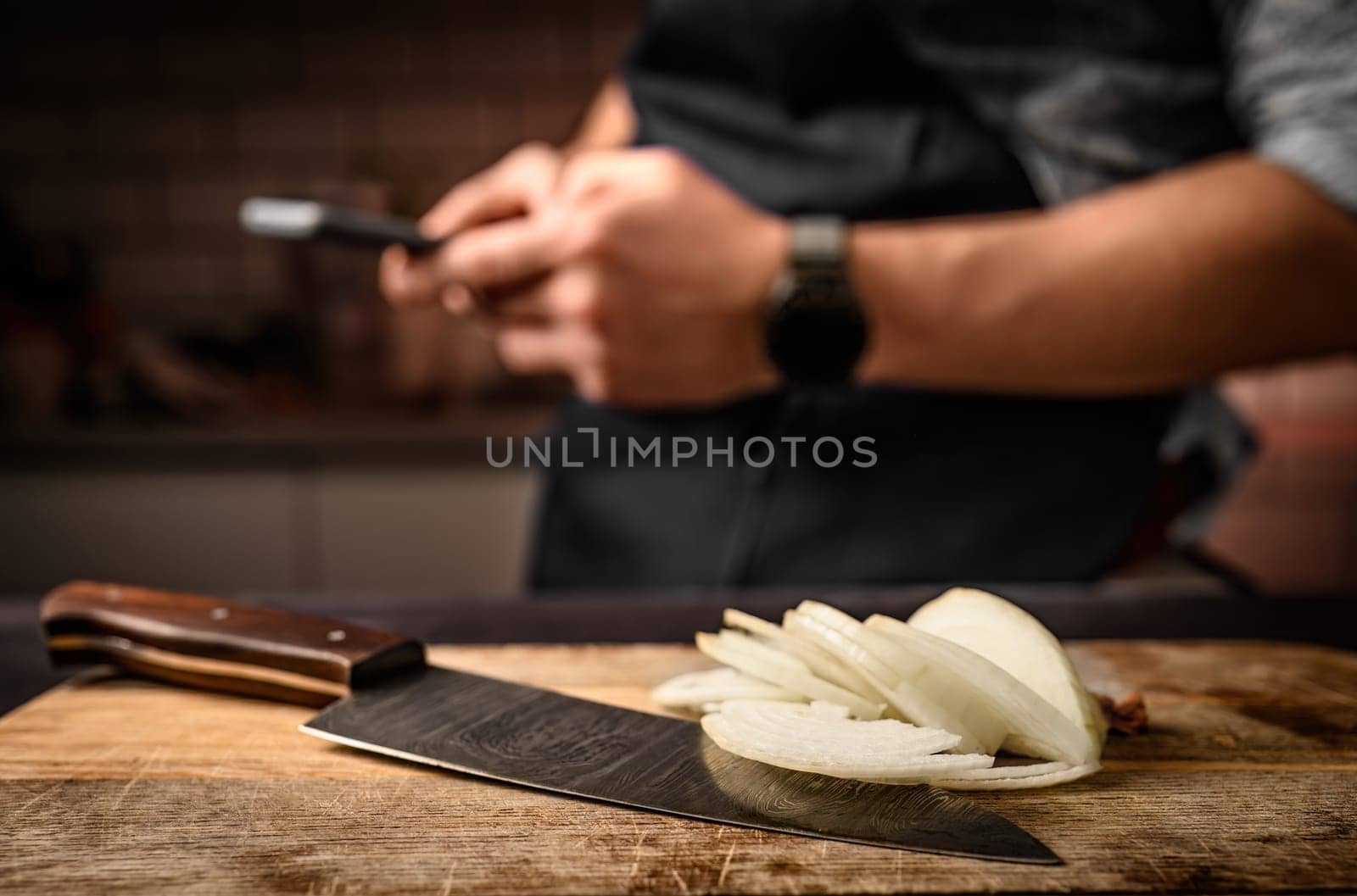 Chief hands holding smartphone at kitchen and sliced onion on wooden board. Man during cooking break with mobile cell phone and bitter vegetable