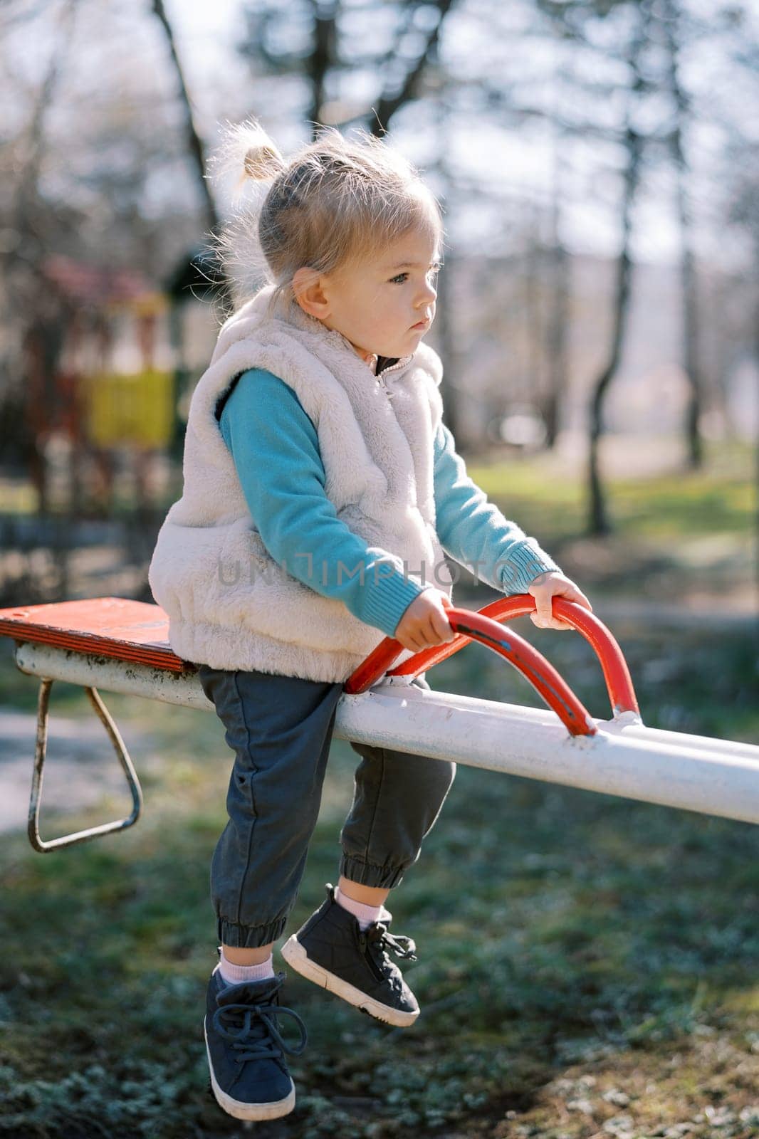 Little girl sits on a metal swing-balancer and holds on to the handles. High quality photo