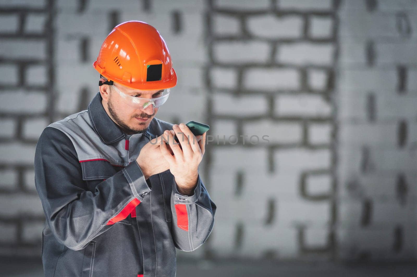 Caucasian male builder in overalls and helmet uses smartphone as radio for communication at construction site.