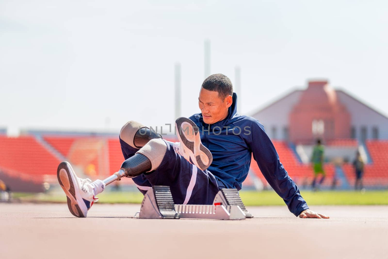 Sport man athlete with a prosthesis on his leg sit on the track and do the stretching of his leg at the stadium with day light.