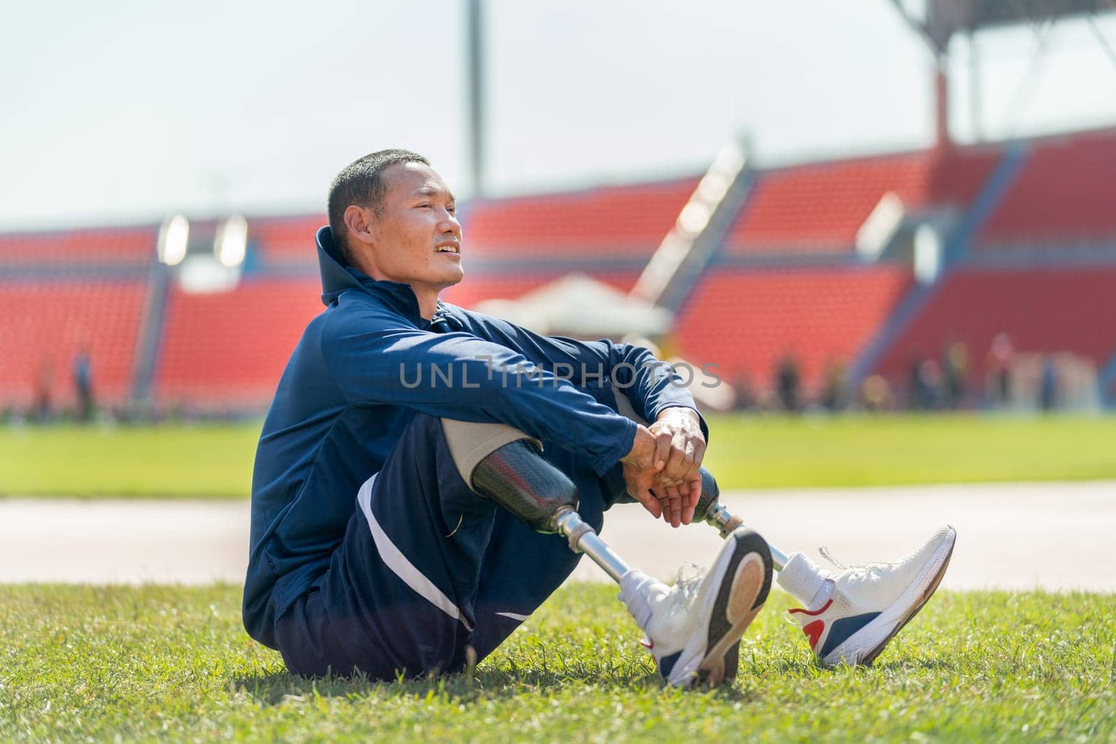 Sport man athlete with a prosthesis on his leg sit on grass field near the track of stadium to relax after exercise with day light.