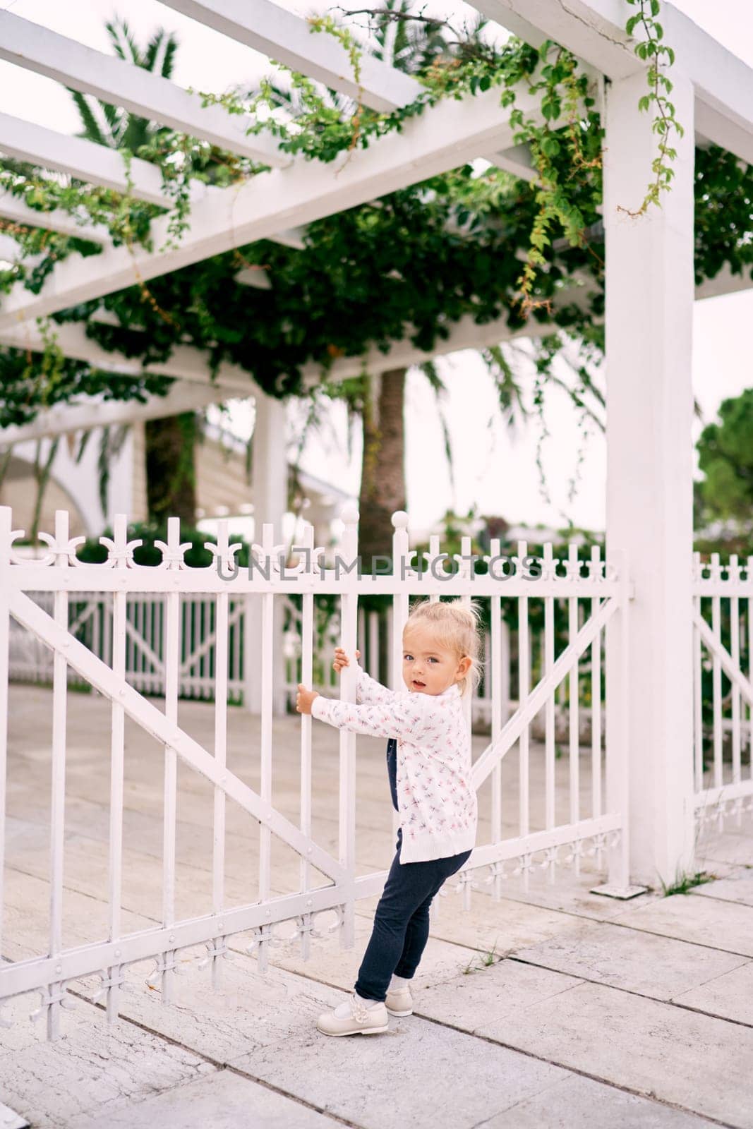 Little girl stands near a pergola fence, holding on to it with her hands and turning her head to the side. High quality photo