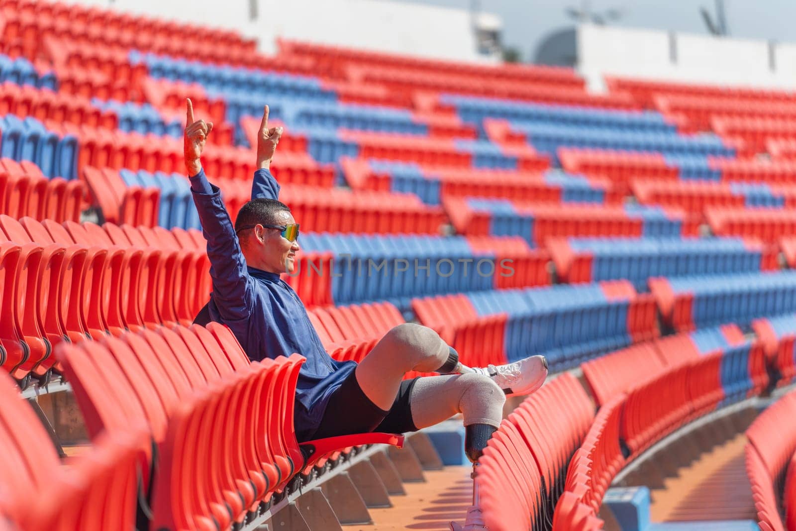 Side view of sport man athlete prosthesis legs sit on chair of amphitheater look forward with show action of happiness by raise two hands up in the stadium with day light.