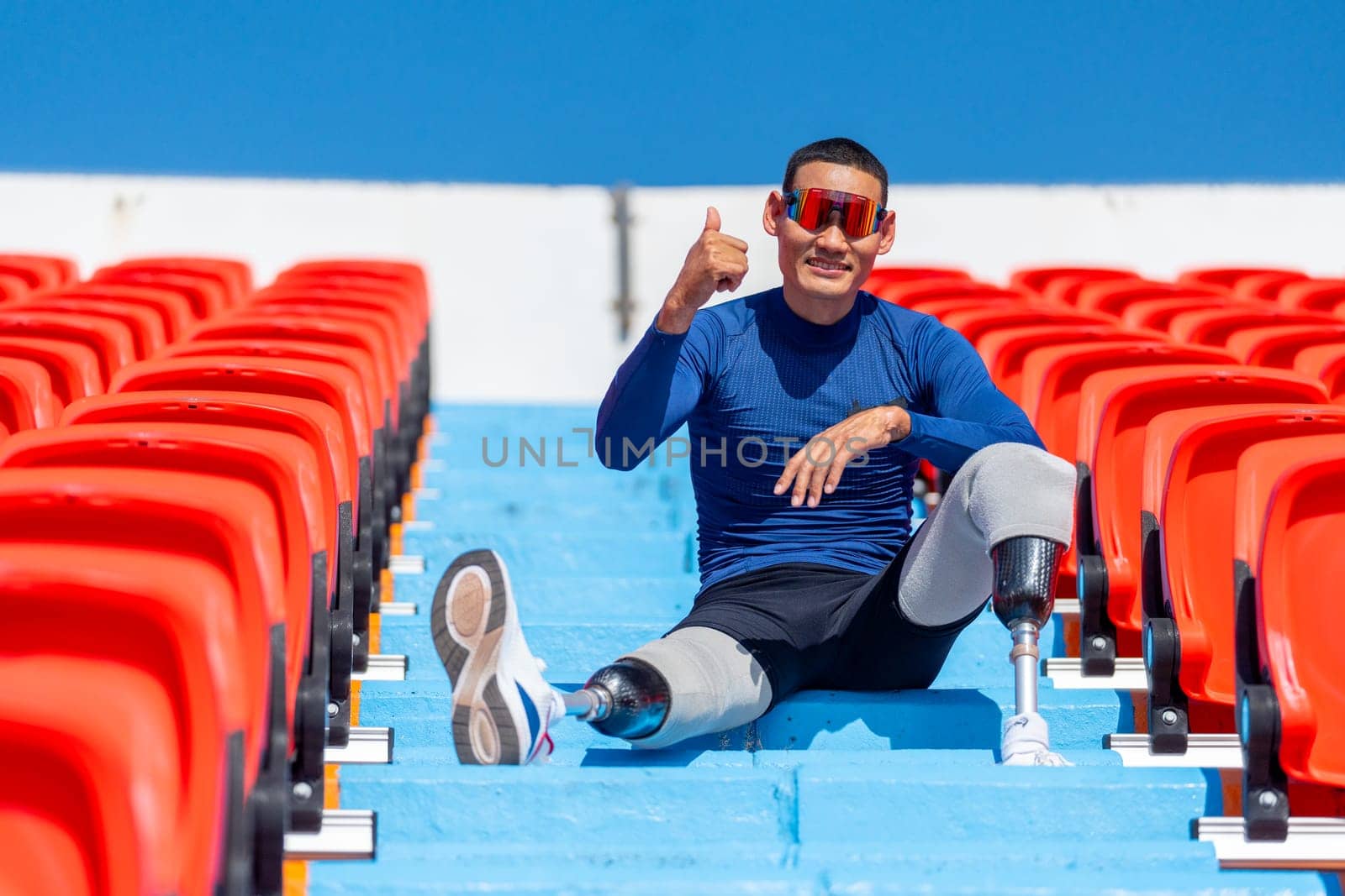 Sport man athlete prosthesis legs sit on stairs with show thumbs up also look at camera on amphitheater in the stadium with day light.