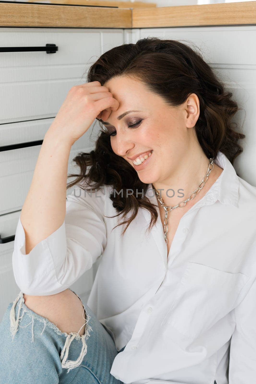 Pretty European emotional laughing woman in a white men's shirt sitting on floor in kitchen. Early morning and smiles when the sun rises. Relaxing and comfortable day.
