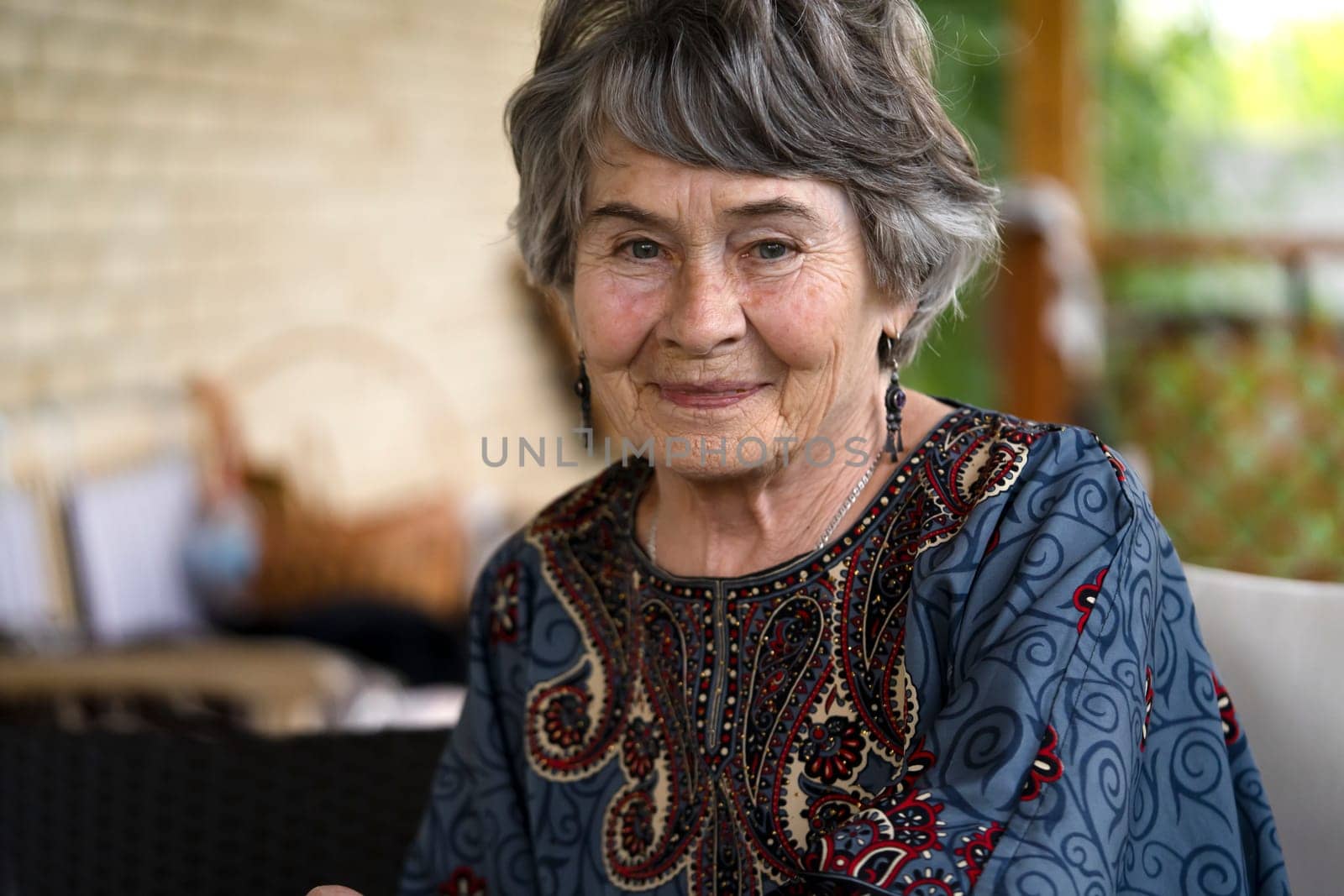 A beautiful elderly woman aged 80+ with a smiling face sits on the veranda of her cozy house in a smart dress, a happy pensioner takes care of her beauty and health.