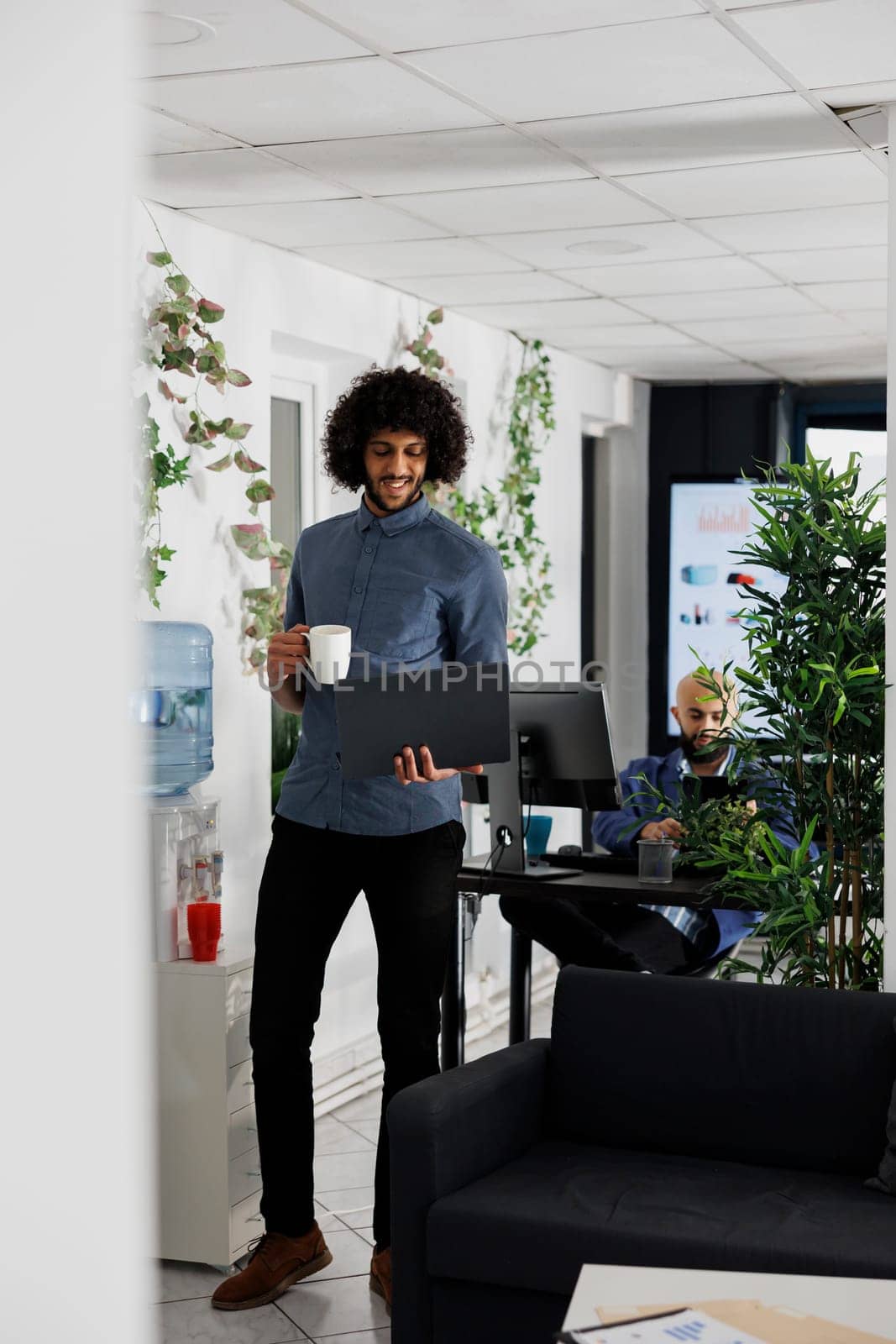 Arab project manager drinking coffee and planning strategy on laptop while standing near workplace table. Young professional holding portable computer and having tea in coworking space