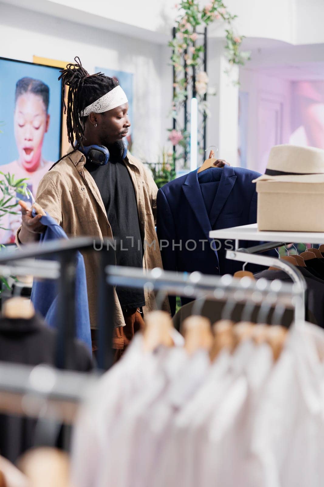 African american man holding two jackets on hangers, examining style and fit. Buyer choosing formal blazer while standing near racks full of clothes and shopping for apparel in mall