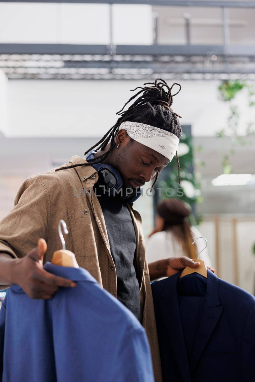 Customer holding two blazers and comparing style in fashion boutique. African american man examining trendy formal jackets on hangers before making purchase in shopping center