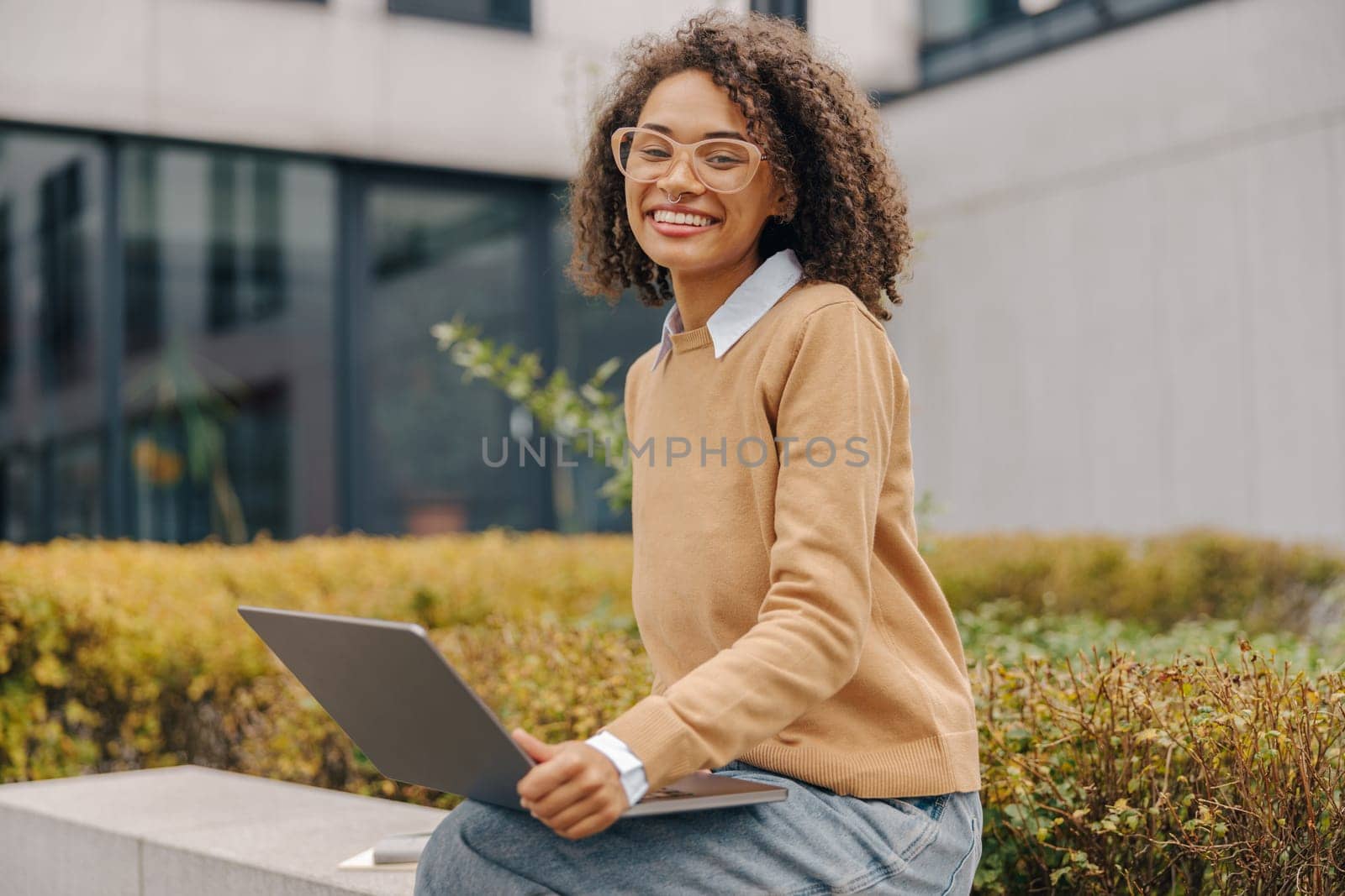 Smart woman student in eyeglasses is using pc laptop sitting outdoors on building background