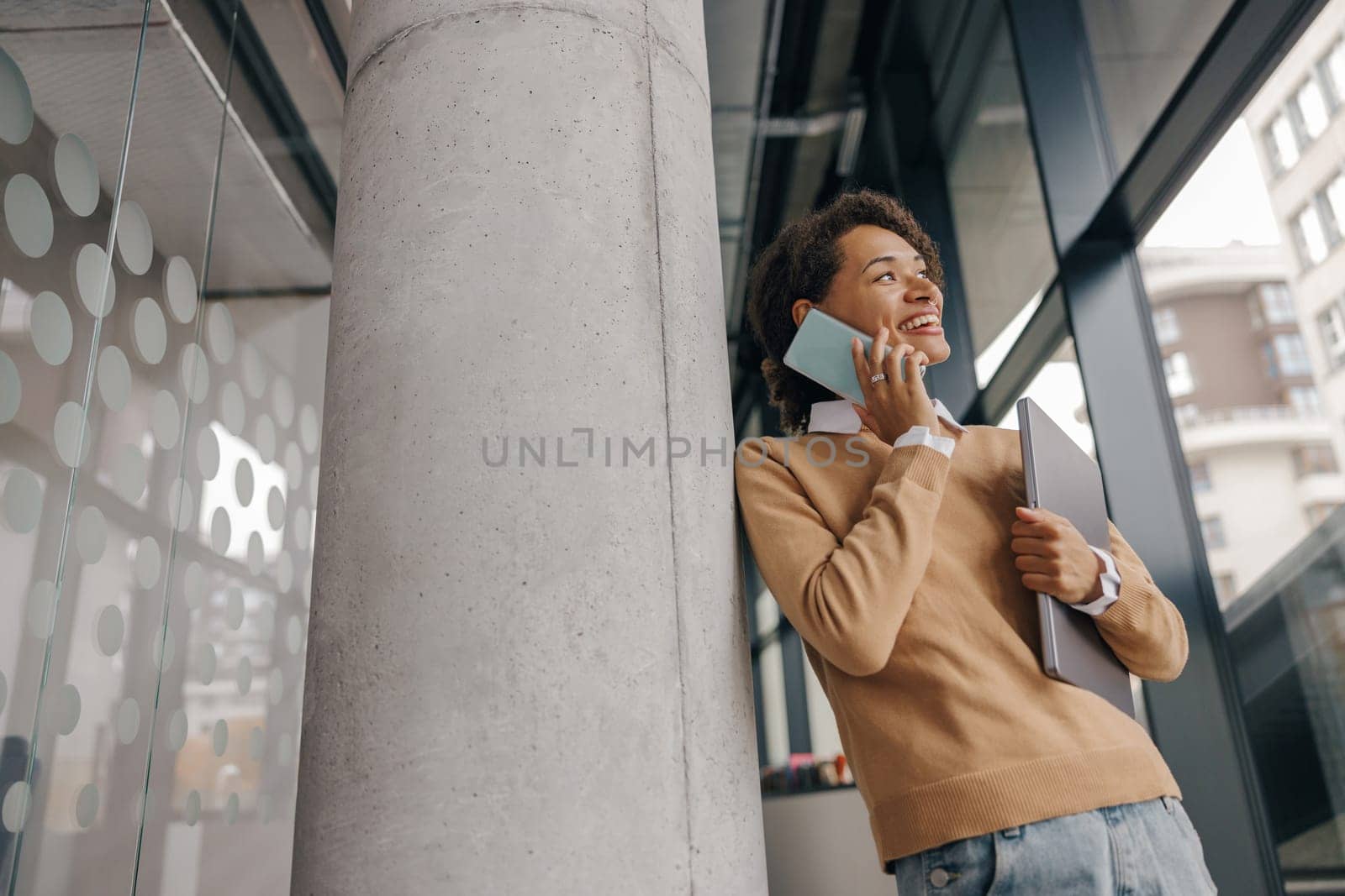 Stylish business woman is talking phone while standing with laptop in modern office