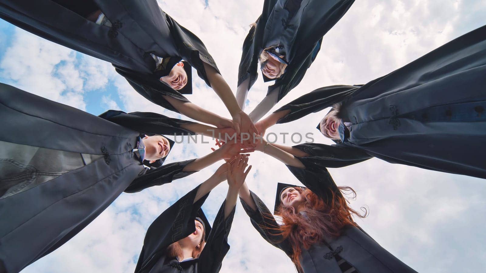 Team of college or university students celebrating graduation. Group of happy successful graduates in academic hats and robes standing in circle and putting their hands together.