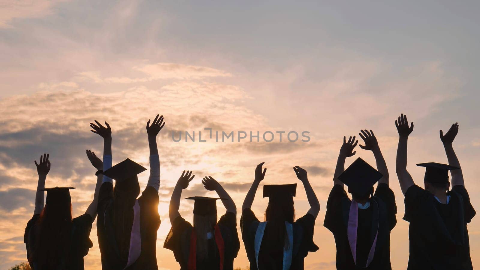 Silhouettes of graduates in black robes waving their arms against the evening sunset