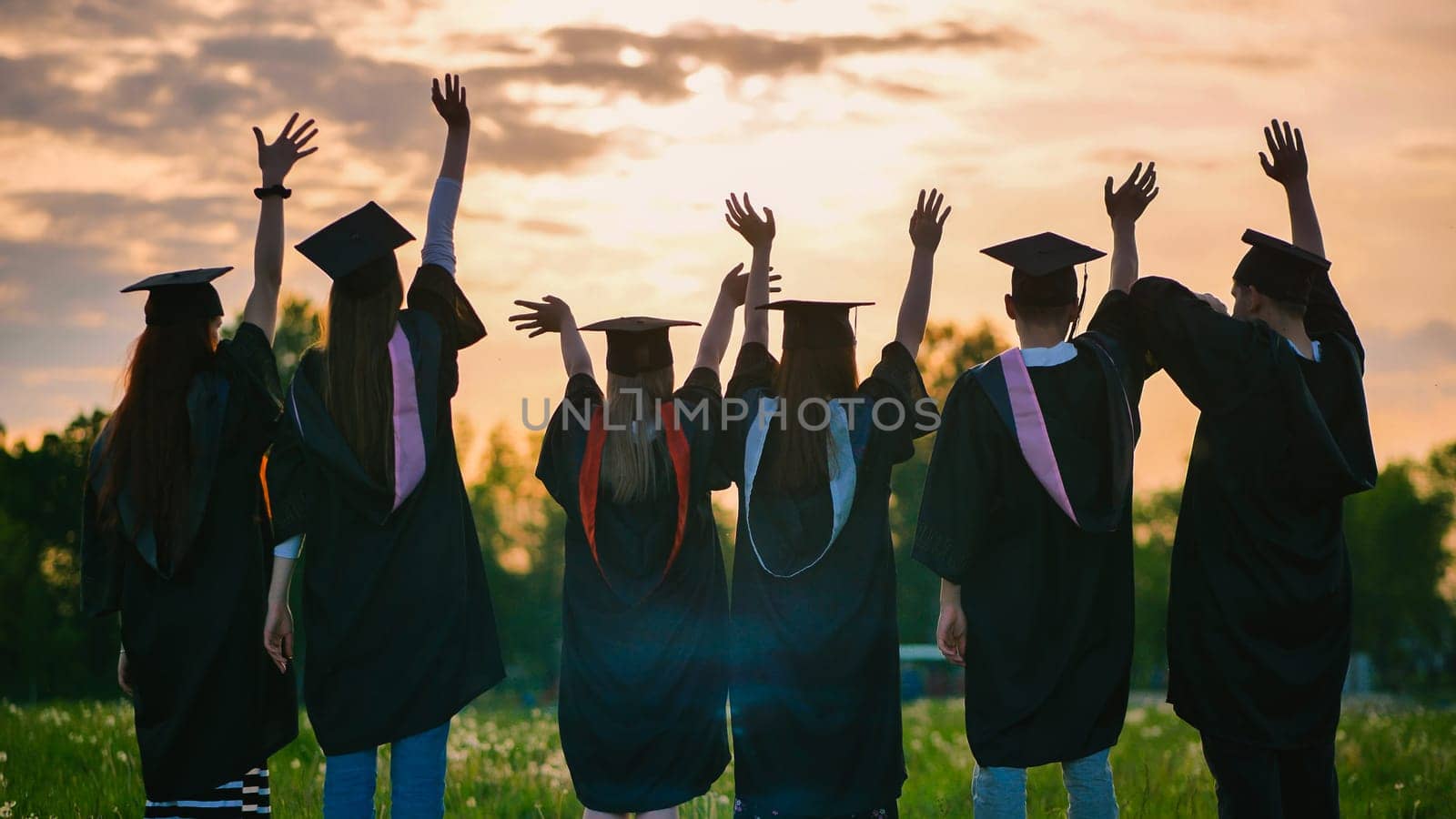 Silhouettes of graduates in black robes waving their arms against the evening sunset. by DovidPro