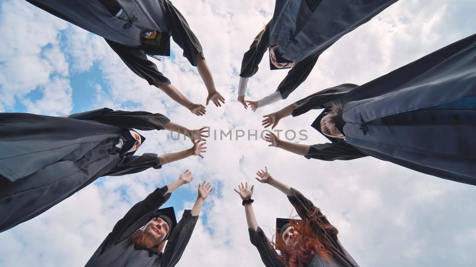 Team of college or university students celebrating graduation. Group of happy successful graduates in academic hats and robes standing in circle and putting their hands together