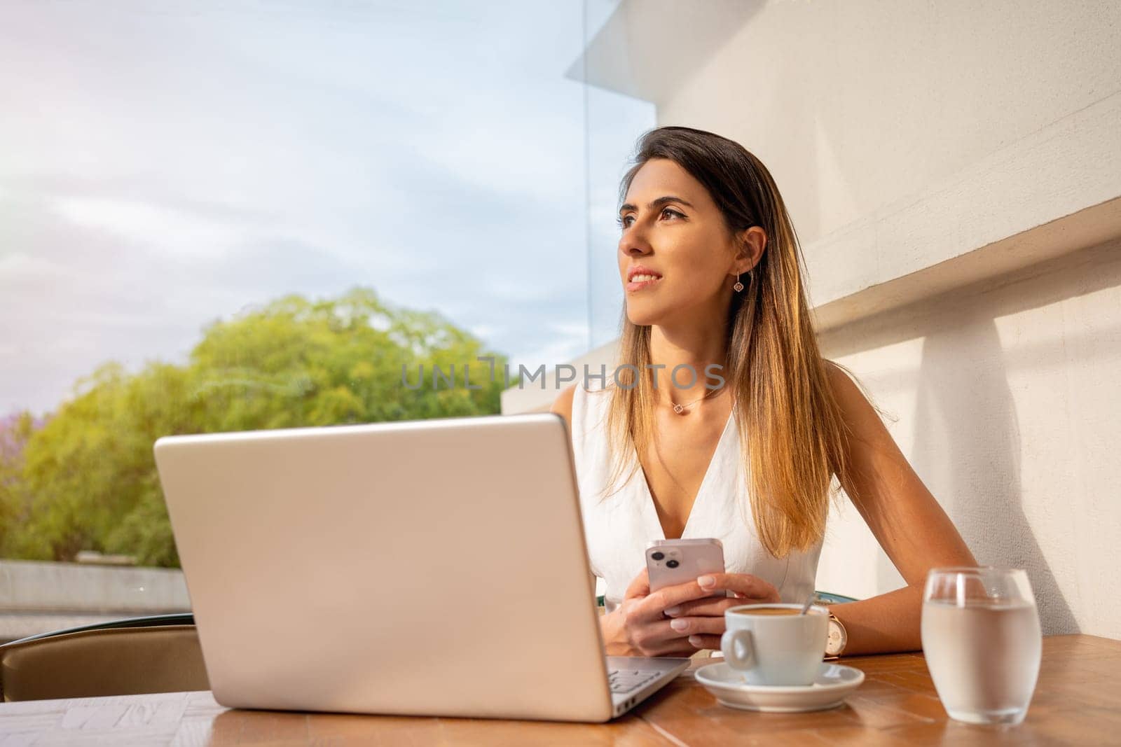 Businesswoman sitting in an outdoor cafe using laptop. Hispanic adult woman sitting in an online meeting in street cafe, drinking coffee. Female freelancer in white dress working or studying on laptop